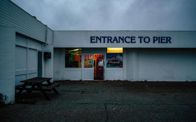 Deserted entrance to Lowestoft South Pier at dusk under a grey sky, an empty picnic bench out front and a single lit doorway glowing inside.
