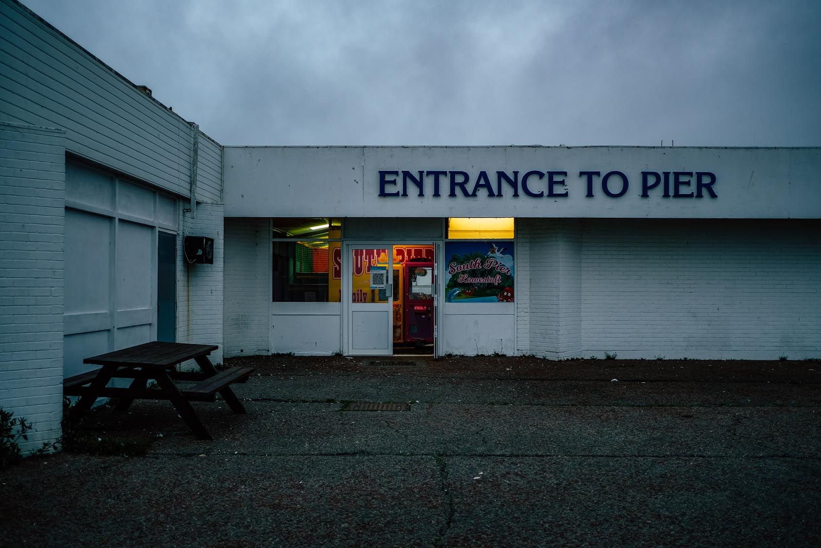 Deserted entrance to Lowestoft South Pier at dusk under a grey sky, an empty picnic bench out front and a single lit doorway glowing inside.