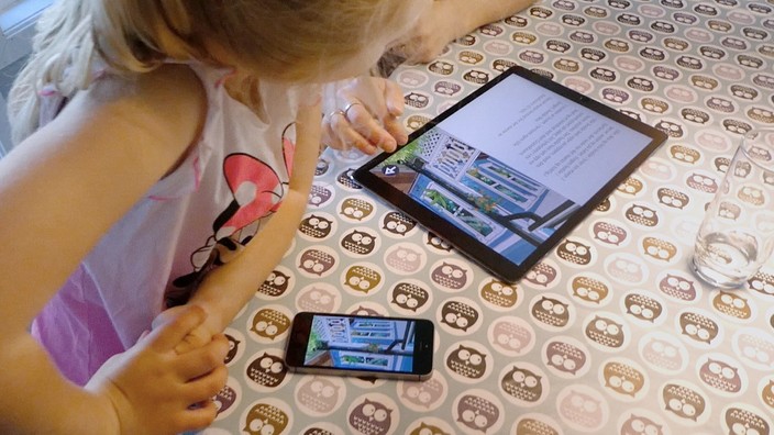 A mother and daughter are using an interactive storybook together. The girl is looking at her mother’s iPad instead of her own iPhone.
