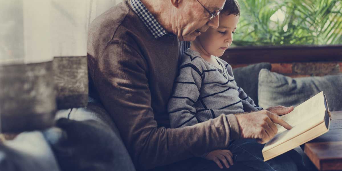 An older man reading a book to a young boy who is sitting on his lap