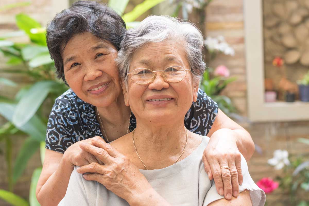 Woman standing behind another woman with her hands on her shoulders. Their faces are next to each other and they are smiling.