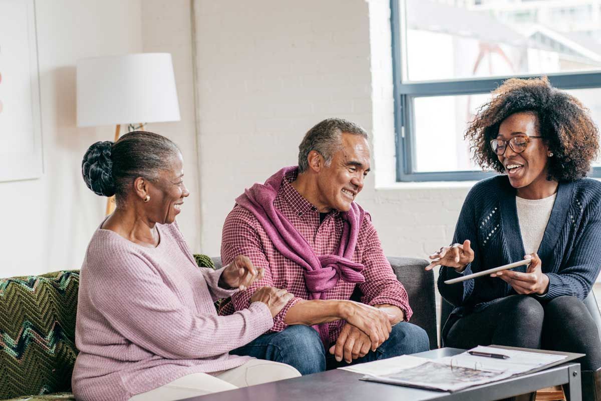 A man and woman sitting on a couch smiling by a woman on the seat next to them who is showing a tablet