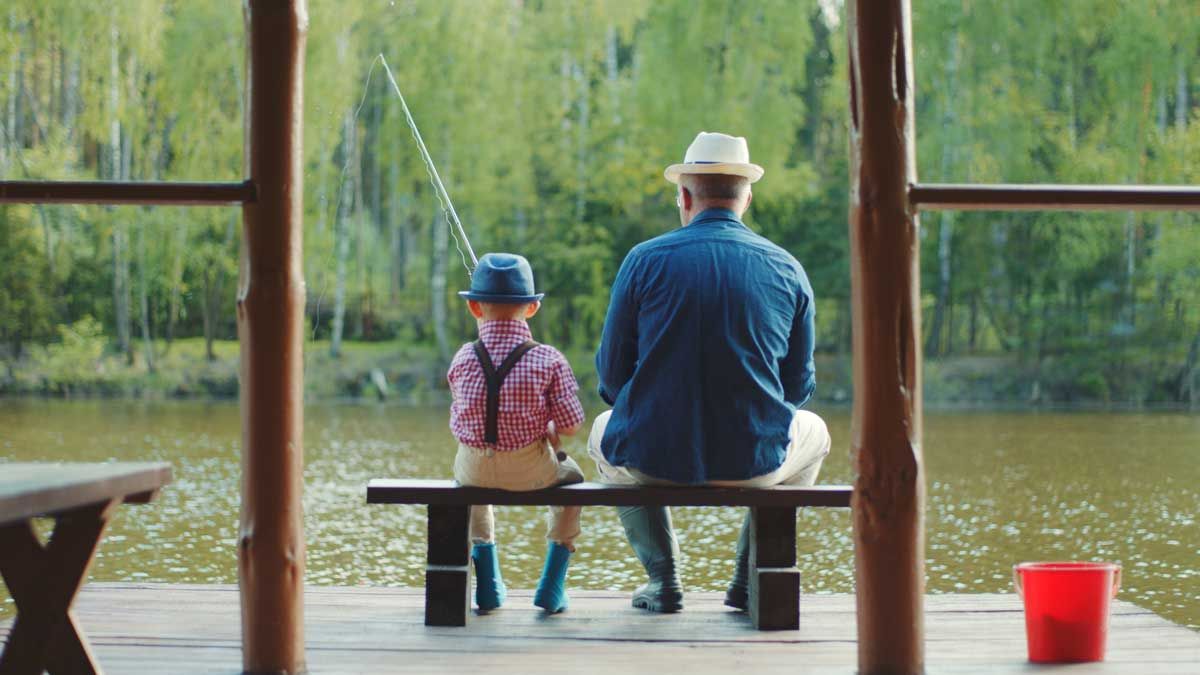 An older man sitting next to a younger boy. We see them from behind. They are fishing.