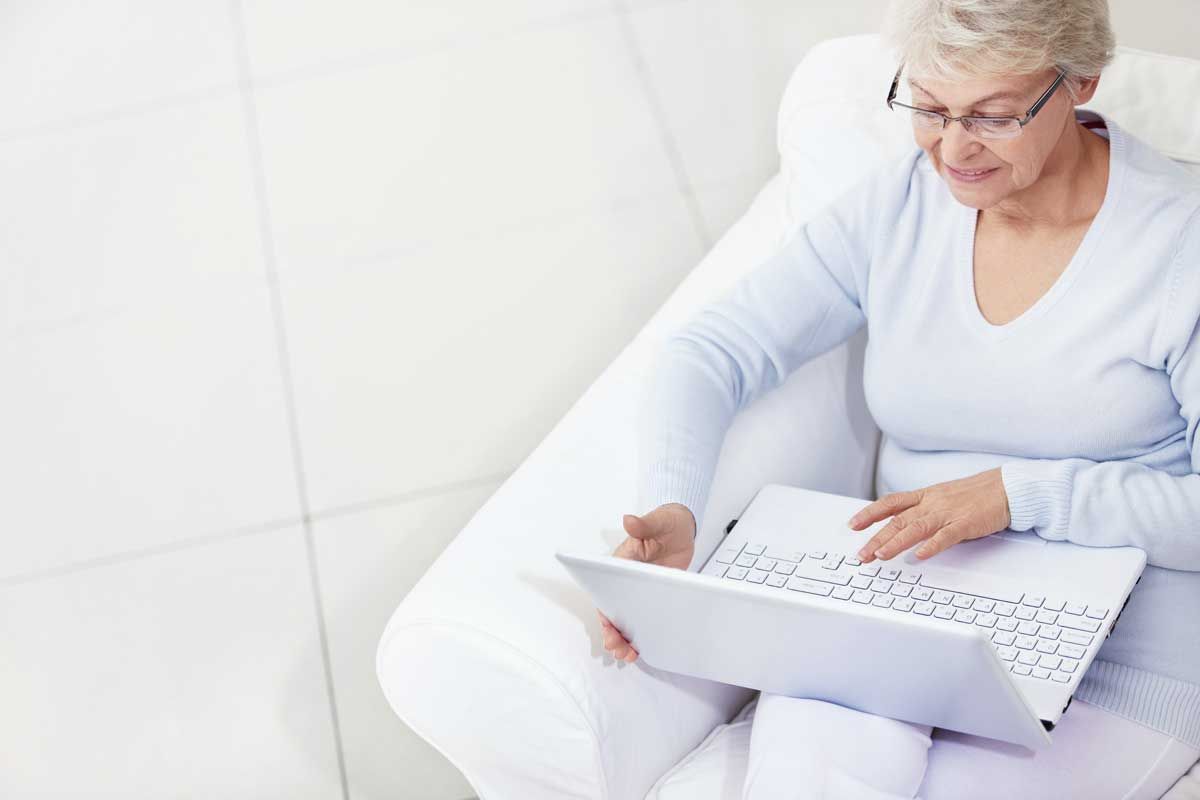 An older woman sitting on a white chair with a white laptop on her lap.