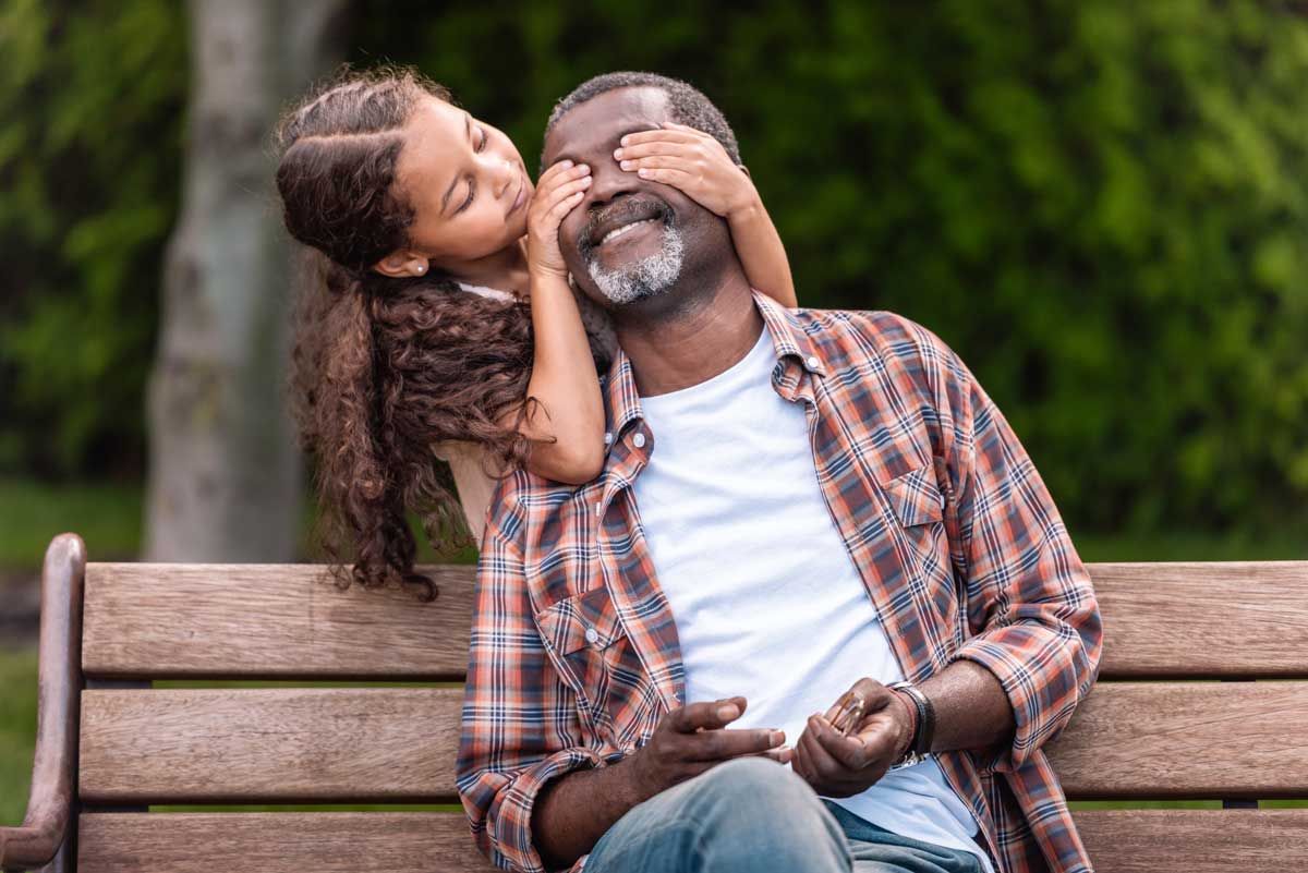 A young girl is standing behind an older man who is sitting on a bench. She is covering his eyes.