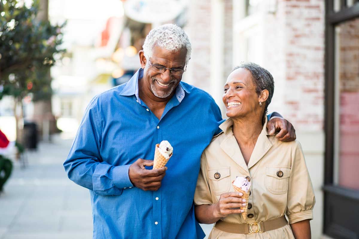 Man and woman walking down the street holding ice cream. The man has is arm around the woman.