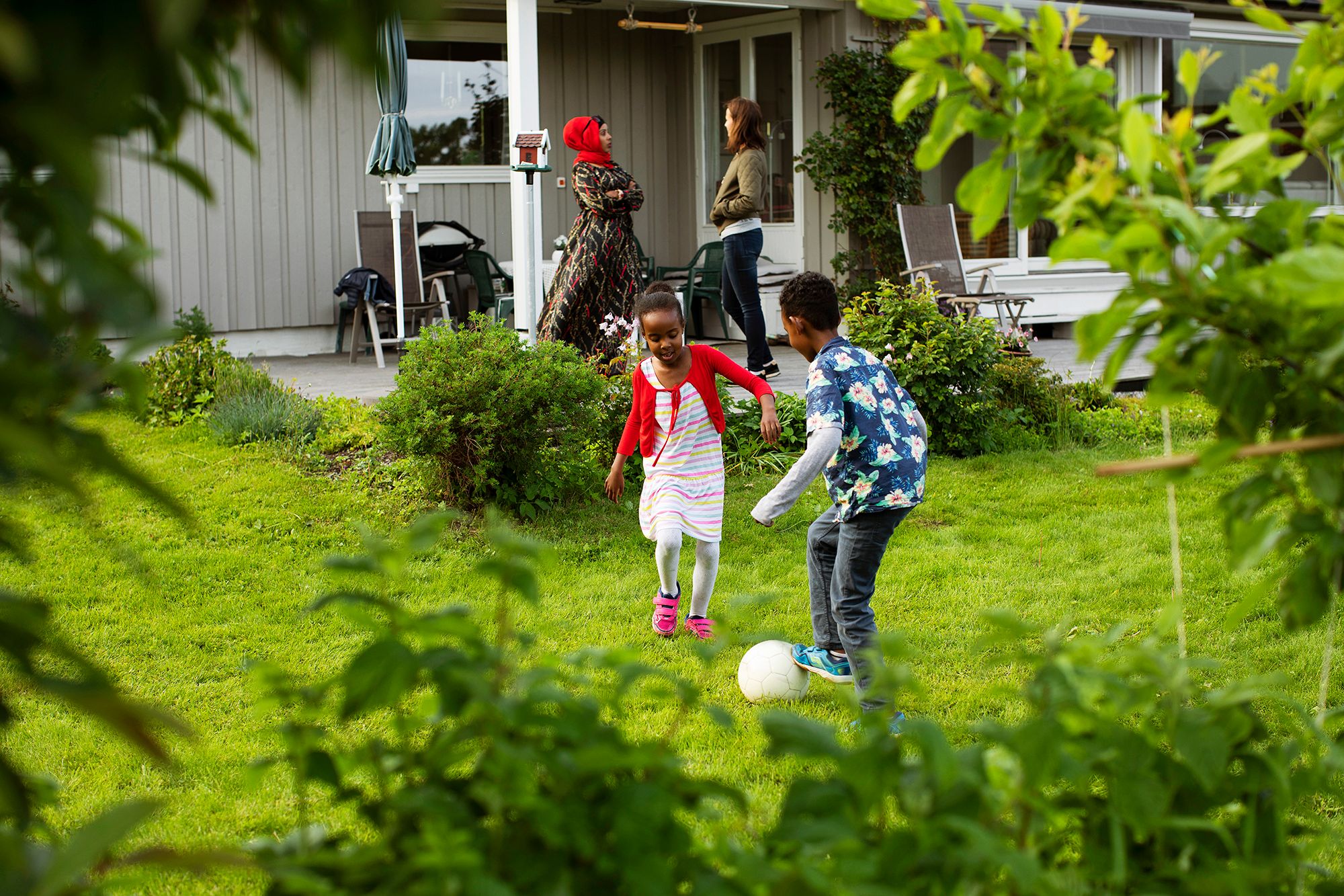 Barn spiller fotball. Mor og familiekontakt i i samtale i bakgrunnen.