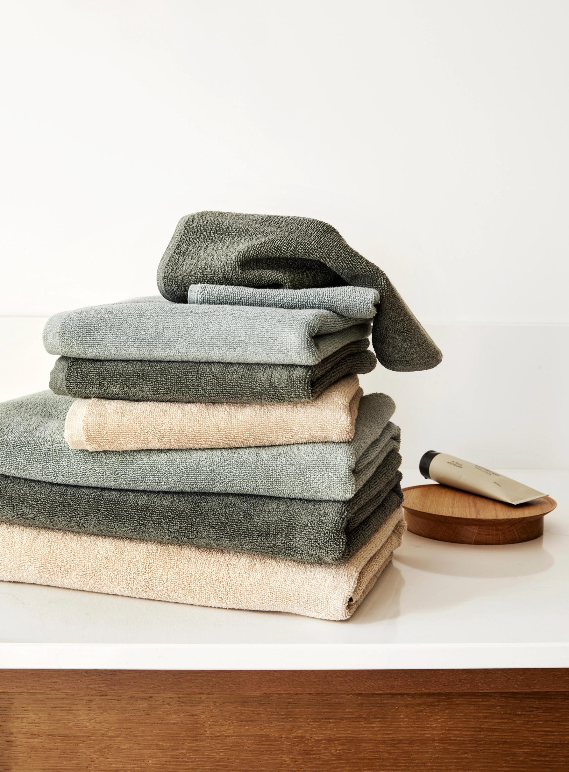 Et minimalistisk oppsett med myke håndklær i ulike jordnære nyanser – olivengrønn, beige, okergul, mørk grå og dyp blå – drapert over en benk i lyst tre. En naturlig kroppsbørste ligger blant de sammenrullede håndklærne, og settingen gir et avslappet og innbydende spa- eller baderomspreg.