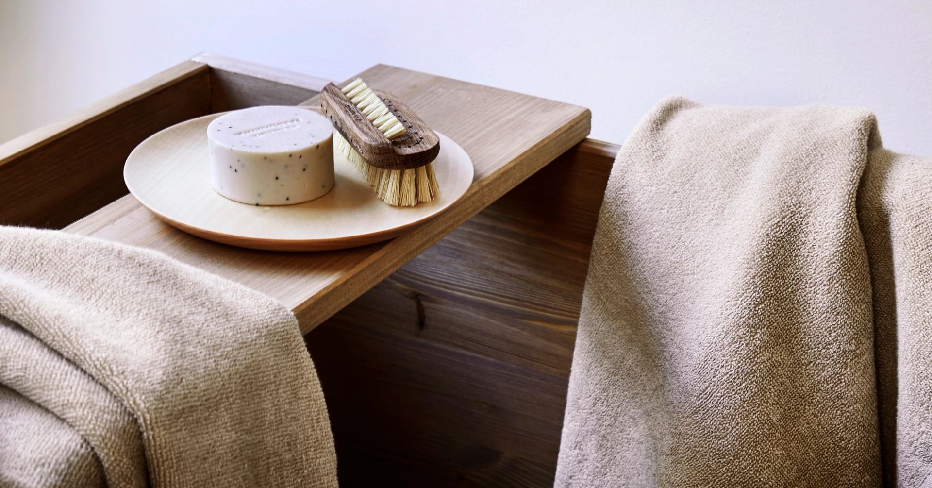 To lyse, beige håndklær i myk frotté ligger drapert over en trekantet benk i lyst treverk. På benken står et rundt fat med en naturlig såpe og en kroppsbørste i tre og naturbust. Bildet formidler en rolig og spa-inspirert stemning med naturlige materialer og varme nøytrale toner.