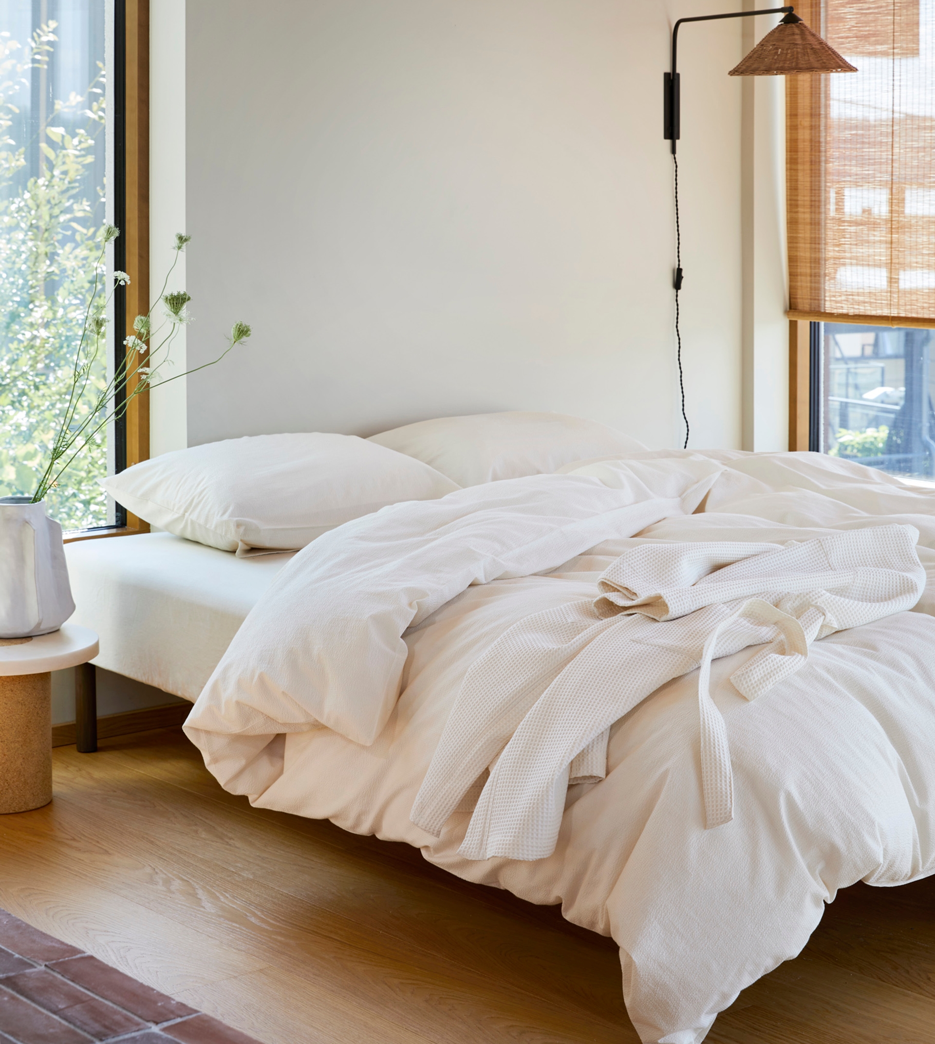 Oppredd seng med Høie Ara sengesett i ubleket bomullskrepp – en naturlig, avslappet stil med teksturert overflate og en kremfarget morgenkåpe dandert over dynen, i et lyst rom med store vinduer og varme trematerialer.