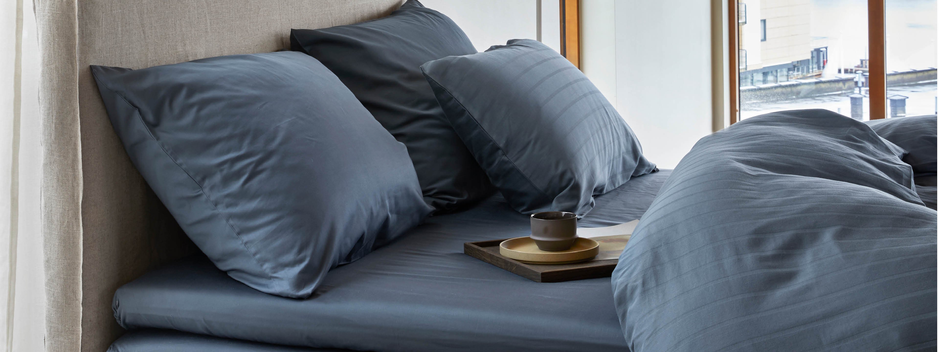 Elegant seng oppredd med glatt, blått satengsengetøy i et moderne rom med store vinduer som gir utsikt over en vannkant og nærliggende bygninger. Sengetøyet har en diskré stripet tekstur som gir et sofistikert preg. På sengen står et lite serveringsbrett med en kopp, som skaper en innbydende og avslappende atmosfære.