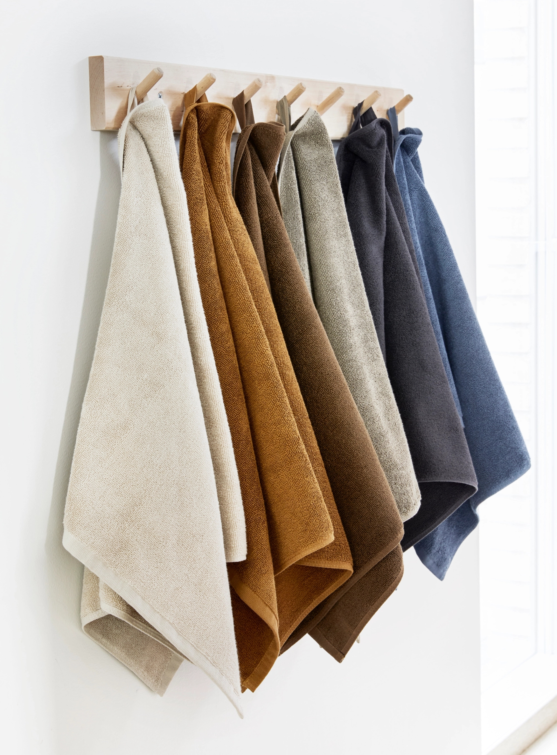 Håndklær i seks ulike farger – ecru, oker, brun, olivengrønn, antrasitt og blå – henger pent på en veggmontert knaggrekke i lyst treverk. Bildet er tatt i naturlig lys og viser en stilren og harmonisk fargepalett for baderommet.
