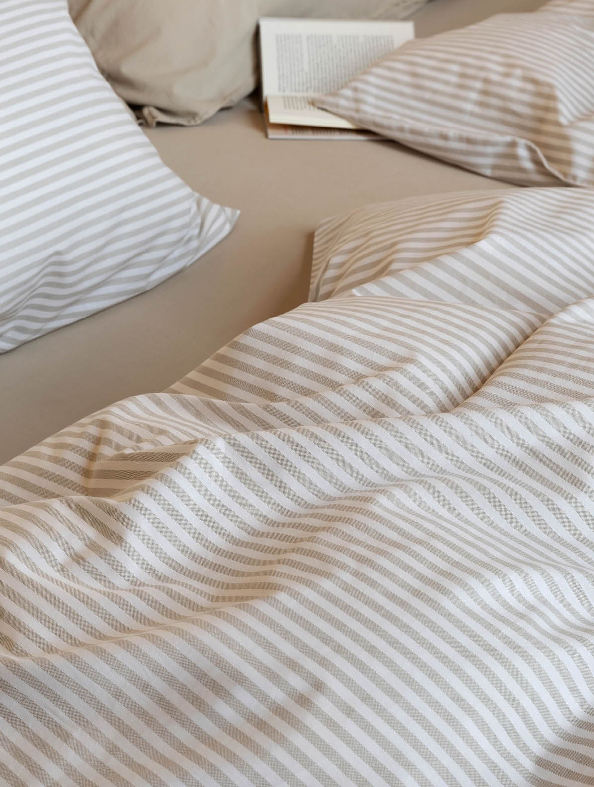 Detalj av oppredd seng med Høie Tor sengesett i beige – smale striper i myk bomull kombinert med ensfarget hotellputetrekk og laken, stylet med naturlige materialer og duse fargetoner for en rolig og enkel soveromsstil.