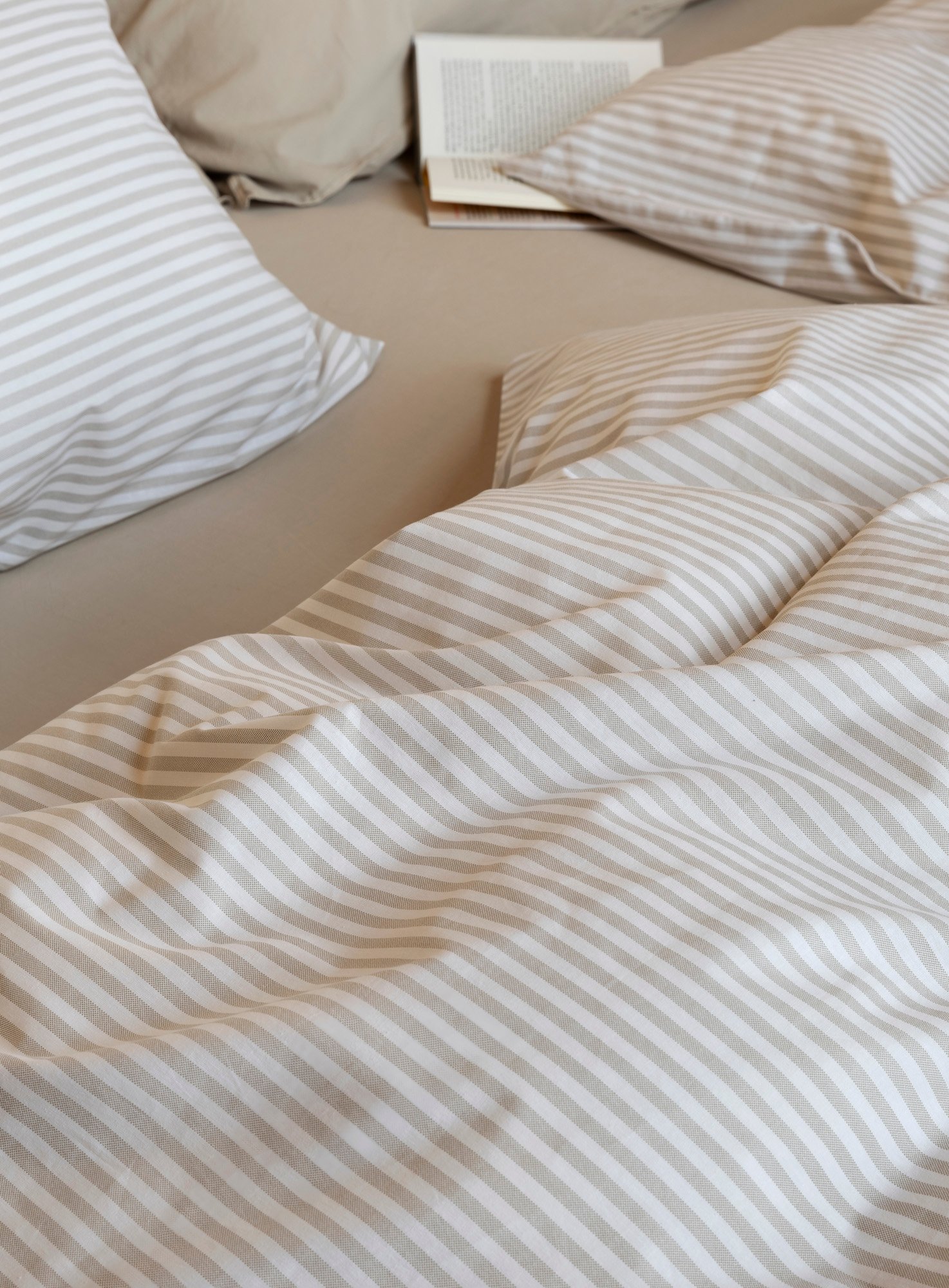 Nærbilde av oppredd seng med Høie Tor og Frøya sengesett i beige – en avslappet miks av smale striper og ensfarget bomull, som skaper en rolig og lun atmosfære på soverommet.