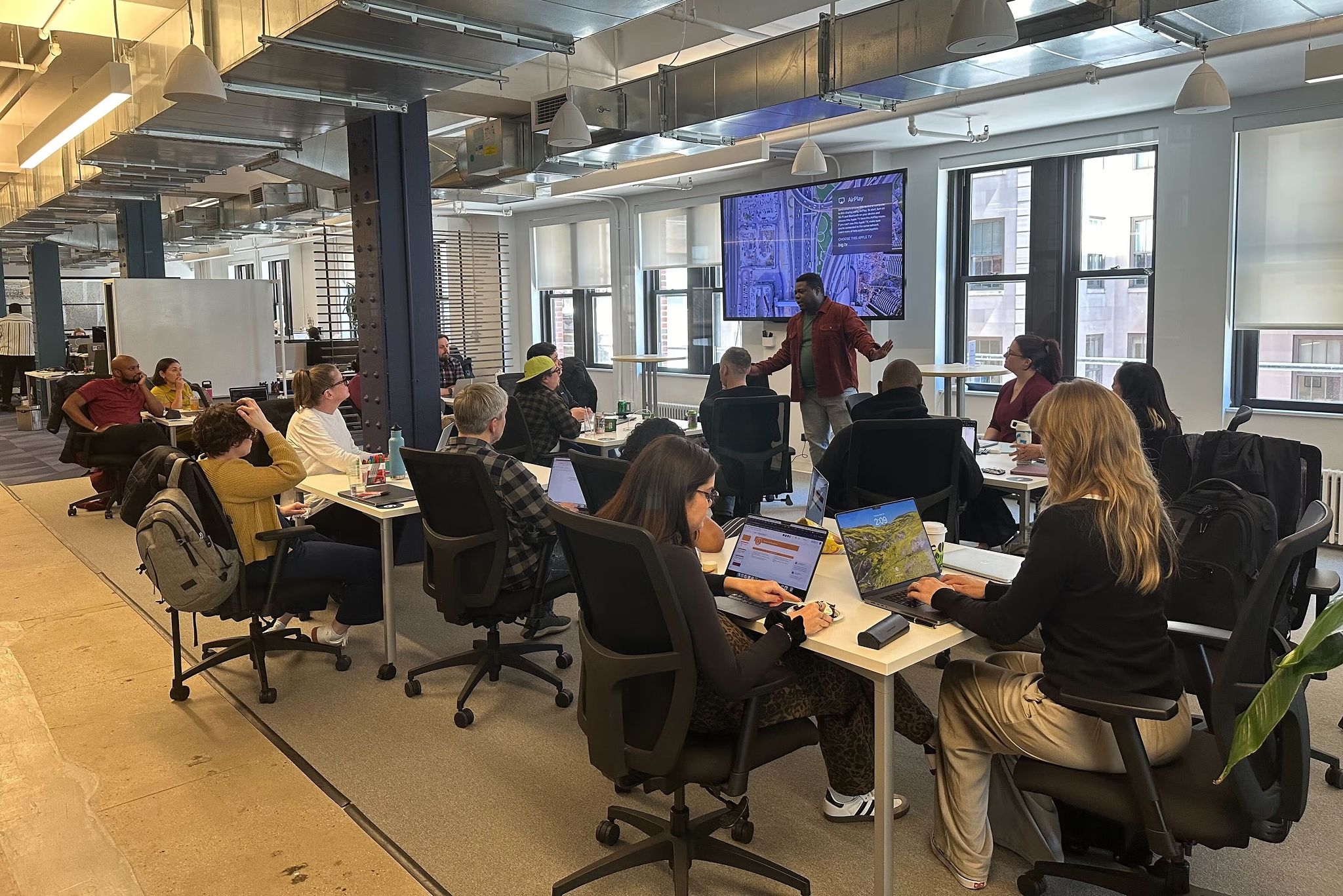 14 teammates sit in a conference space listening to one person standing in front of a large digital screen.