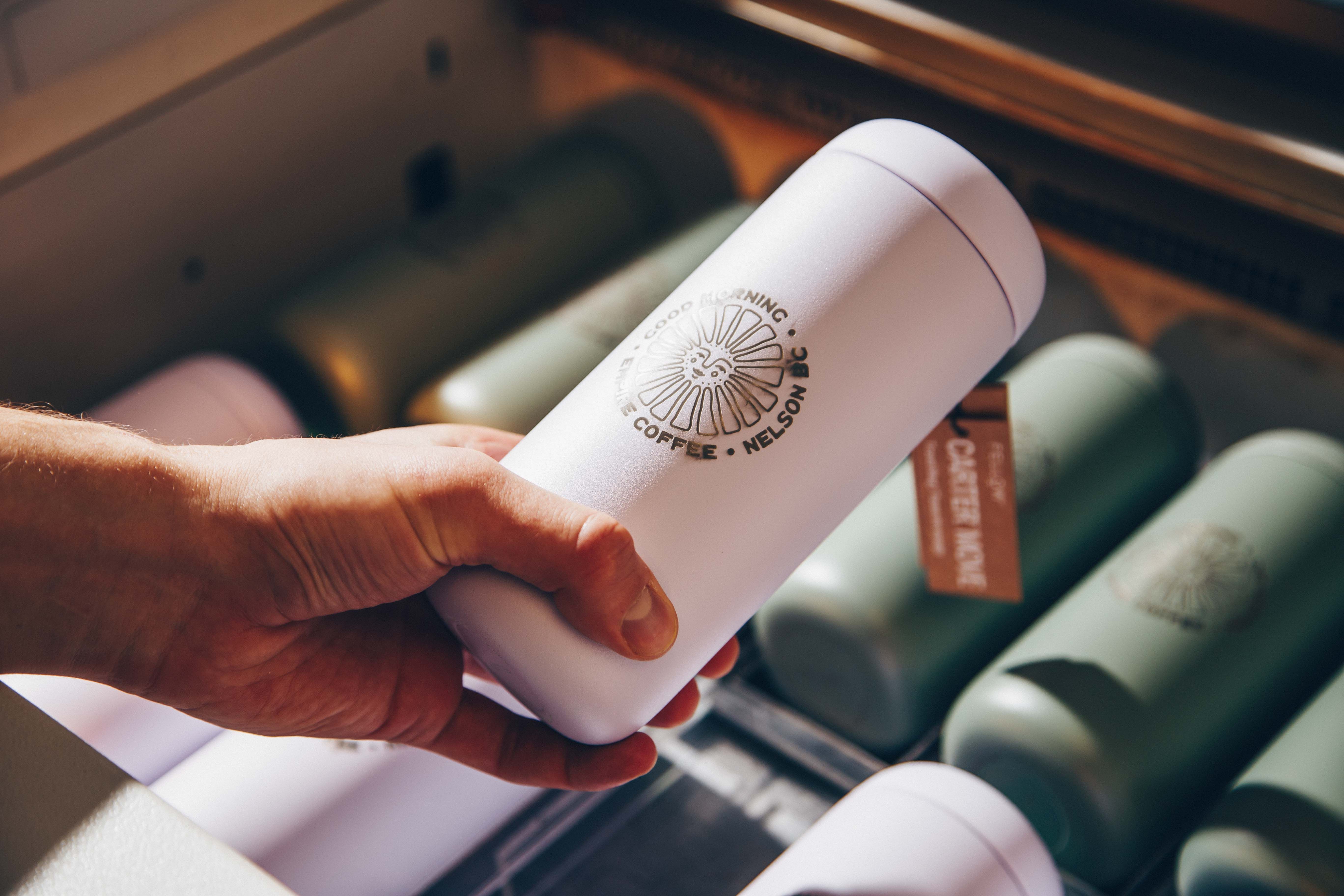 A hand holding a custom-engraved white tumbler with the logo "Good Morning Coffee, Nelson BC." The background shows a collection of similar tumblers, emphasizing personalized drinkware for branding or promotional use.