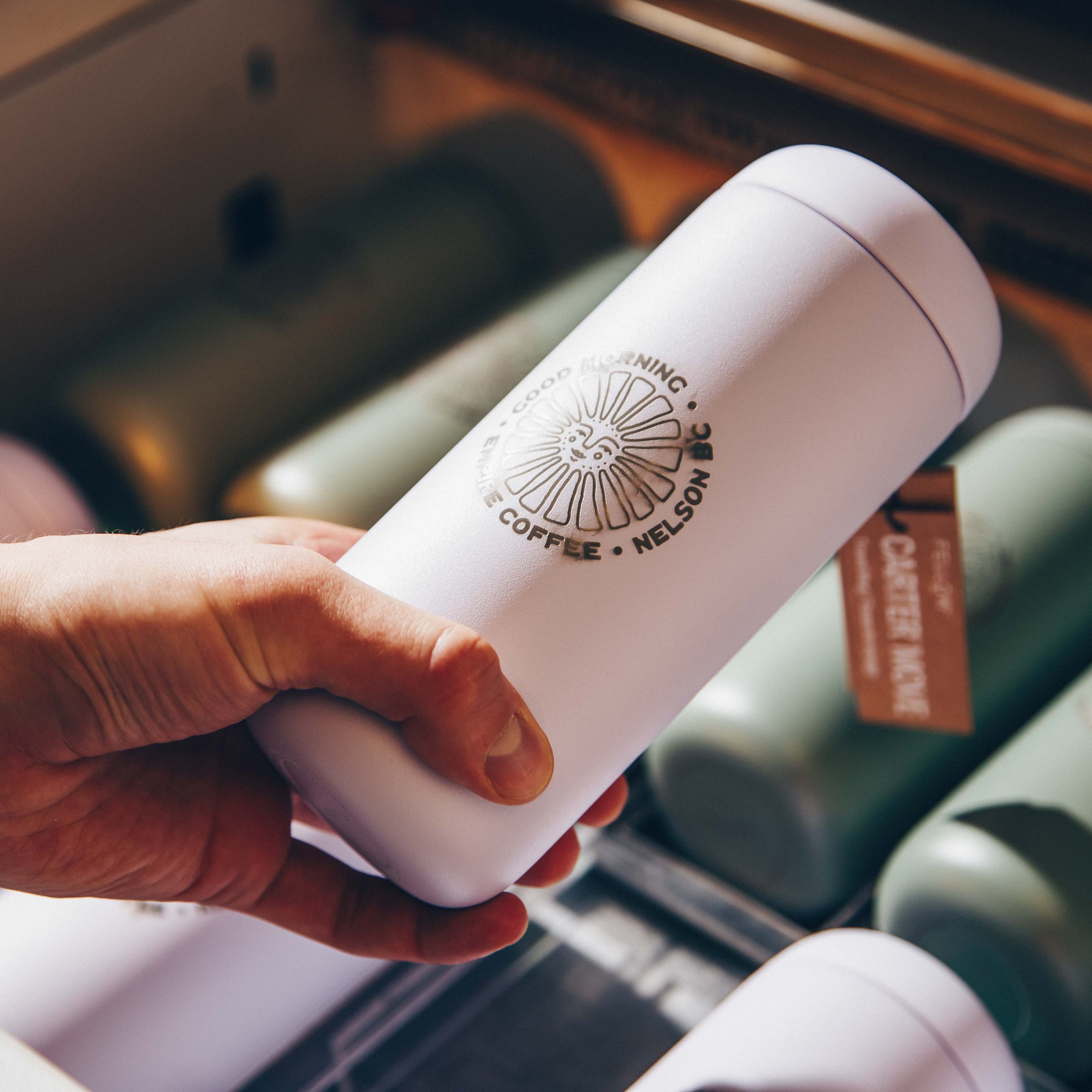 A hand holding a custom-engraved white tumbler with the logo "Good Morning Coffee, Nelson BC." The background shows a collection of similar tumblers, emphasizing personalized drinkware for branding or promotional use.