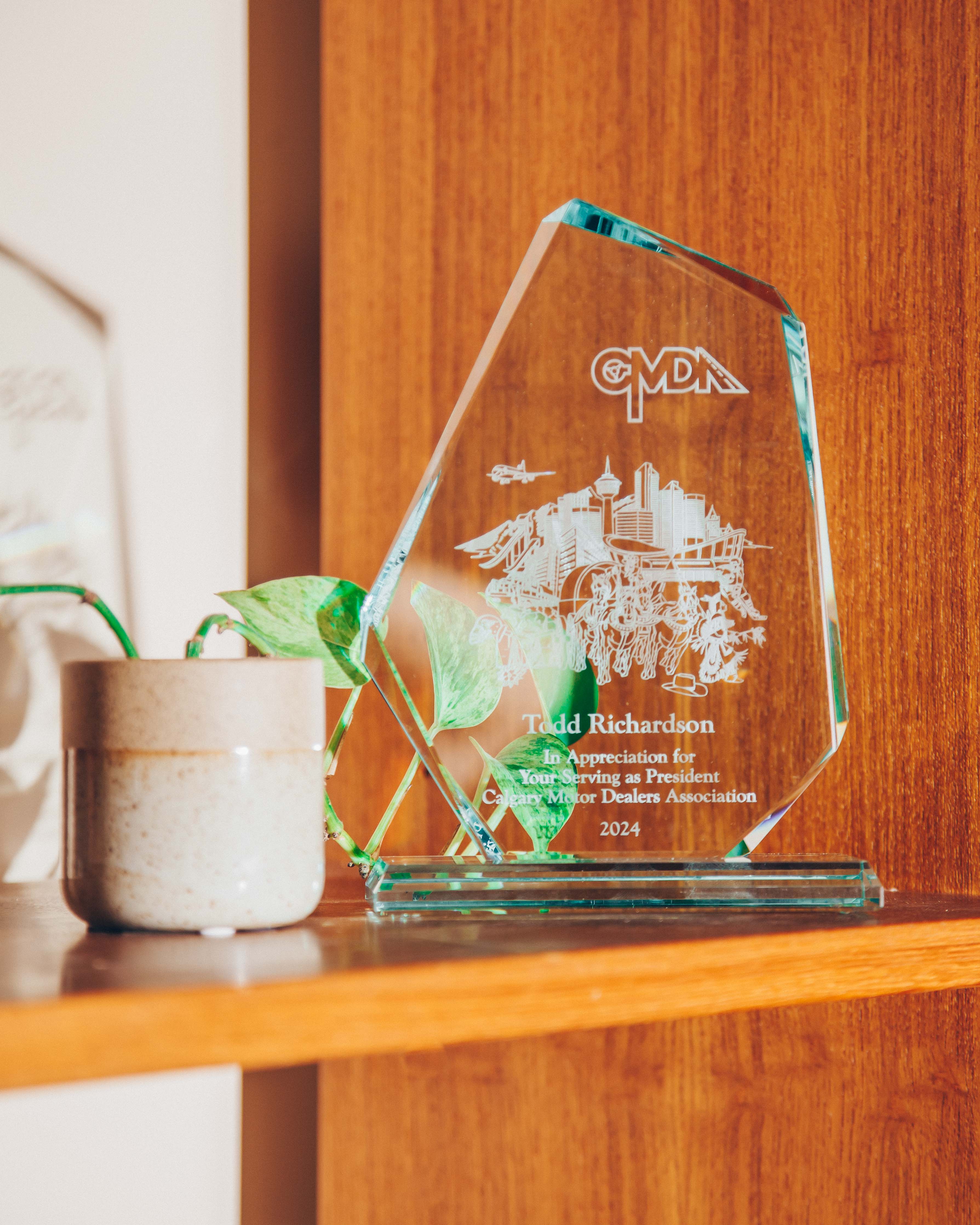 A glass award placed on a wooden shelf. The award is clear with engraved text and images, including a cityscape and the letters 'CMDA' at the top. The text on the award reads 'Todd Richardson, In Appreciation for Your Serving as President, Canadian Motor Dealers Association, 2024.' Next to the award is a small ceramic pot with a green plant, adding a touch of nature to the scene. The background is a warm wooden surface, contributing to a cozy and inviting atmosphere. The lighting is soft and natural, casting gentle shadows and highlighting the textures of the wood and the glass award.