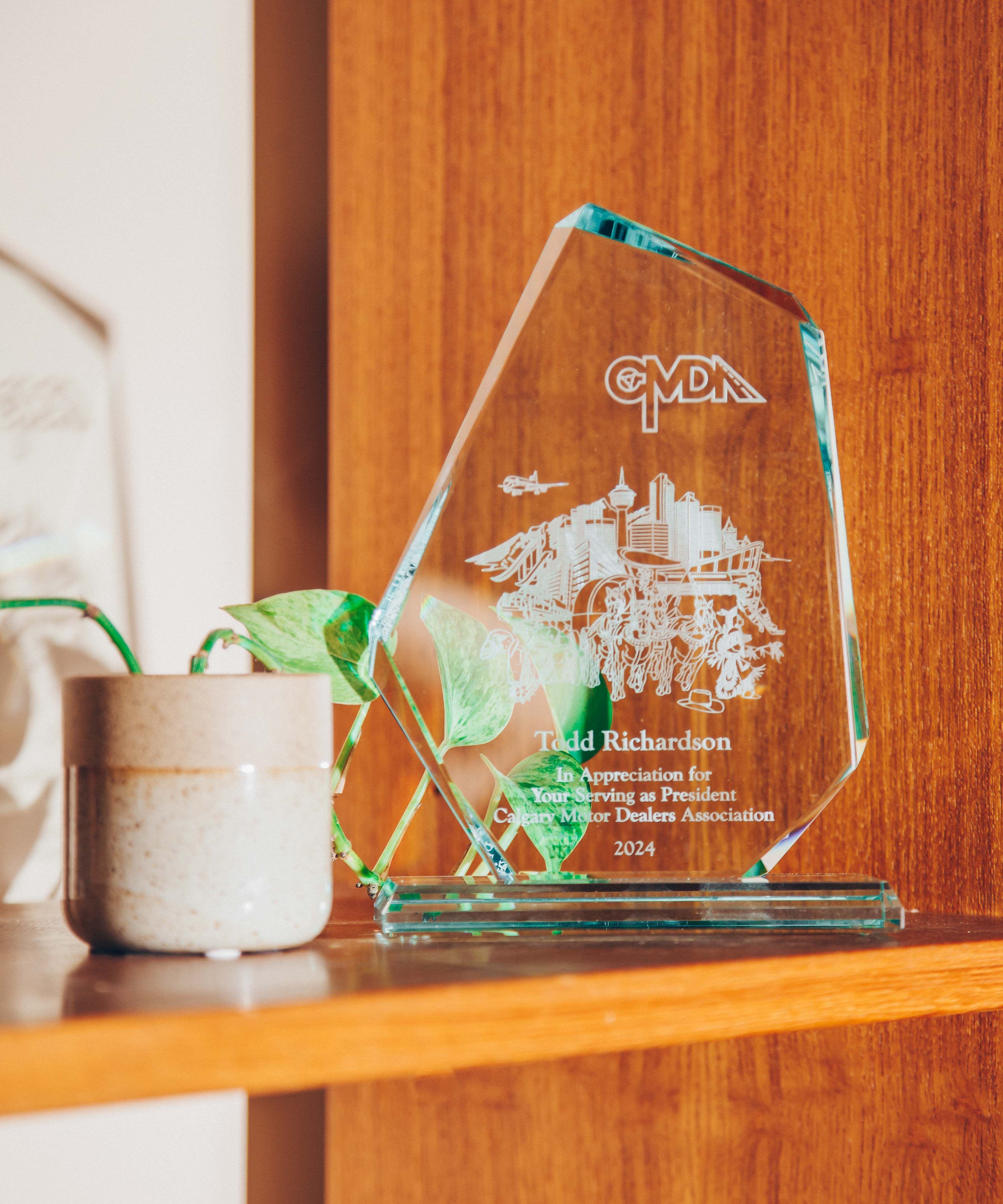 A glass award placed on a wooden shelf. The award is clear with engraved text and images, including a cityscape and the letters 'CMDA' at the top. The text on the award reads 'Todd Richardson, In Appreciation for Your Serving as President, Canadian Motor Dealers Association, 2024.' Next to the award is a small ceramic pot with a green plant, adding a touch of nature to the scene. The background is a warm wooden surface, contributing to a cozy and inviting atmosphere. The lighting is soft and natural, casting gentle shadows and highlighting the textures of the wood and the glass award.