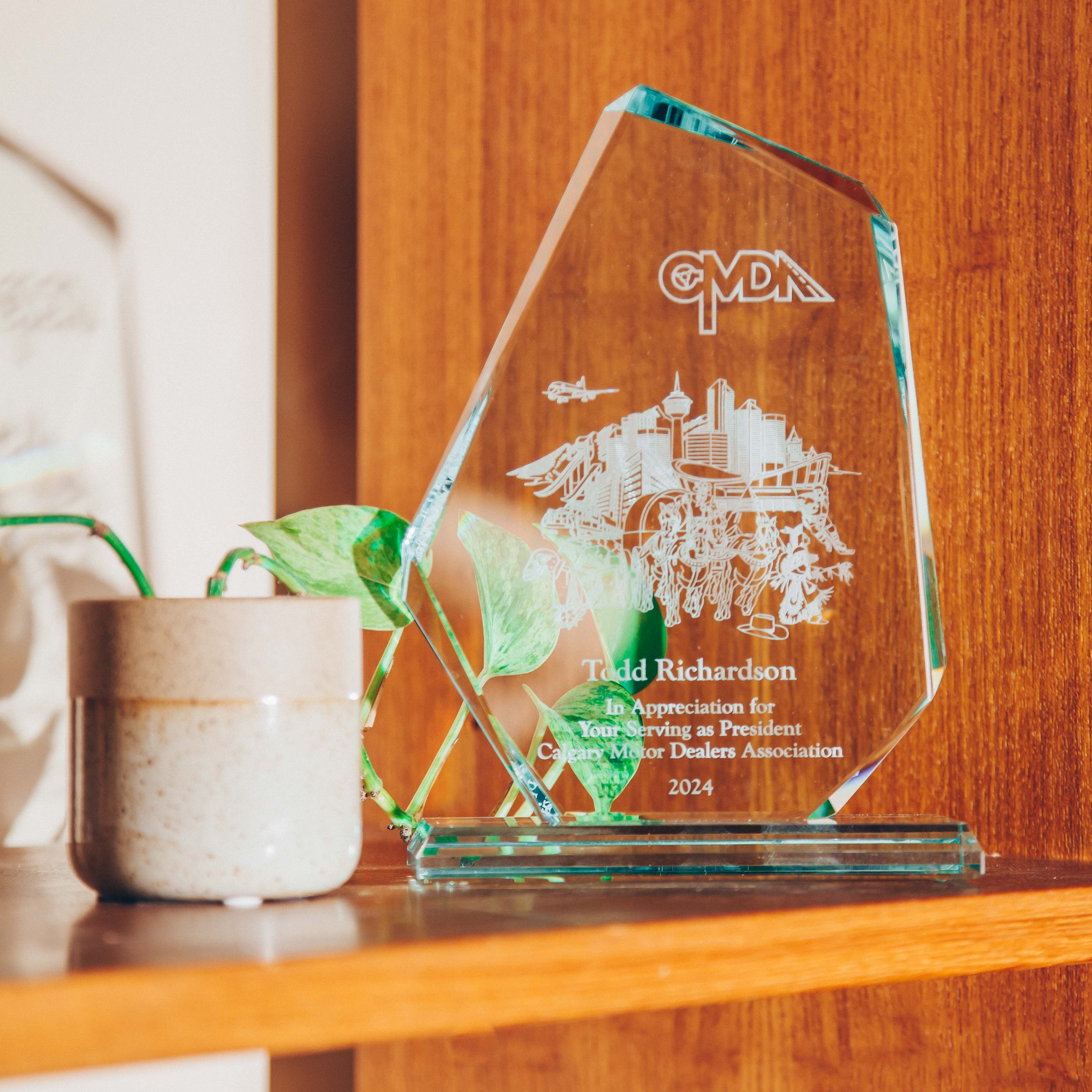 A glass award placed on a wooden shelf. The award is clear with engraved text and images, including a cityscape and the letters 'CMDA' at the top. The text on the award reads 'Todd Richardson, In Appreciation for Your Serving as President, Canadian Motor Dealers Association, 2024.' Next to the award is a small ceramic pot with a green plant, adding a touch of nature to the scene. The background is a warm wooden surface, contributing to a cozy and inviting atmosphere. The lighting is soft and natural, casting gentle shadows and highlighting the textures of the wood and the glass award.
