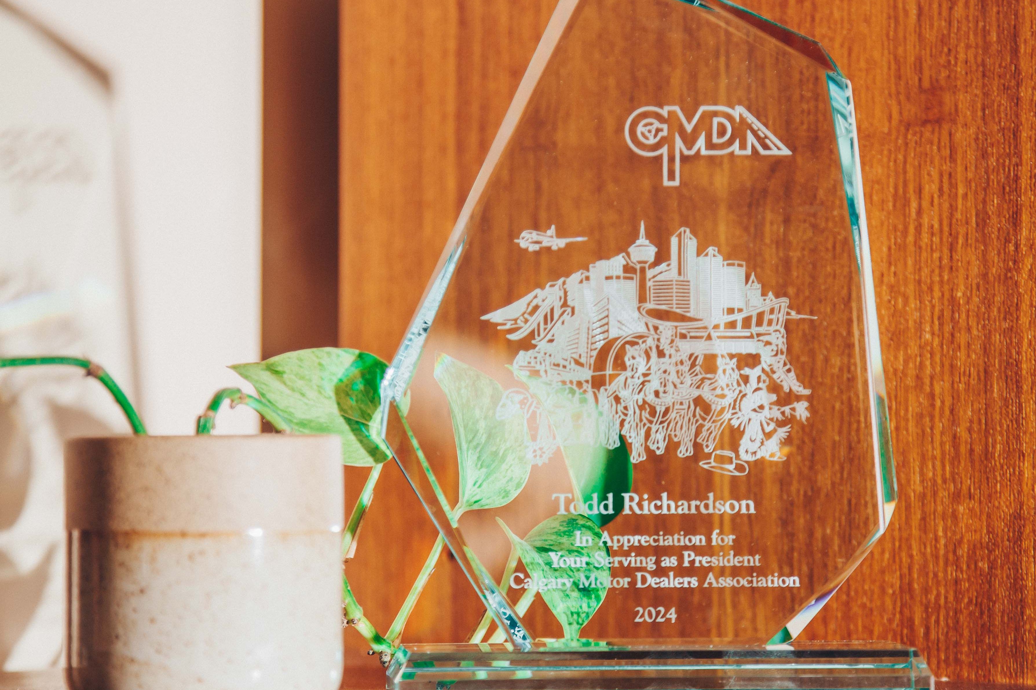 A glass award placed on a wooden shelf. The award is clear with engraved text and images, including a cityscape and the letters 'CMDA' at the top. The text on the award reads 'Todd Richardson, In Appreciation for Your Serving as President, Canadian Motor Dealers Association, 2024.' Next to the award is a small ceramic pot with a green plant, adding a touch of nature to the scene. The background is a warm wooden surface, contributing to a cozy and inviting atmosphere. The lighting is soft and natural, casting gentle shadows and highlighting the textures of the wood and the glass award.