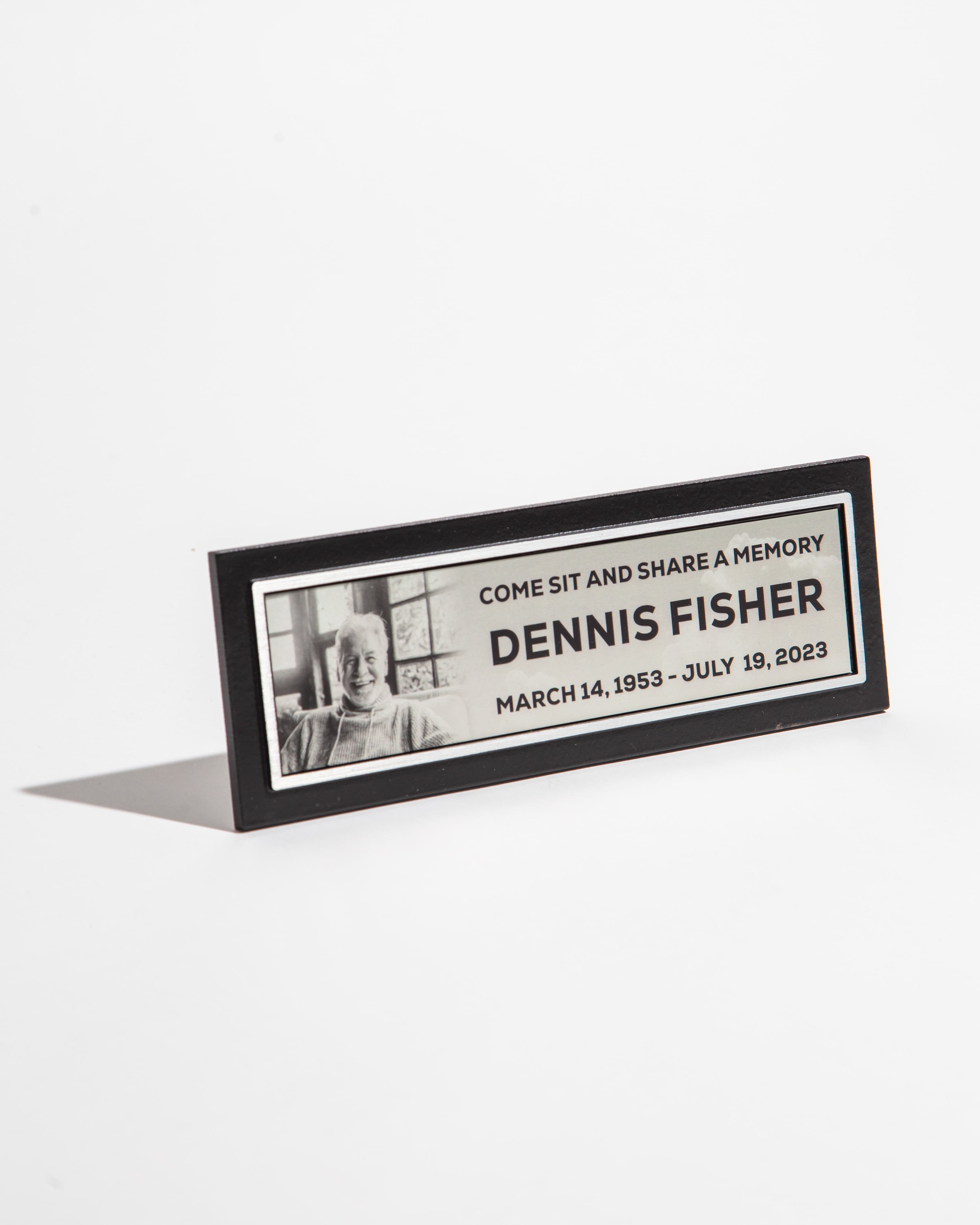 Memorial plaque with a photo and text commemorating Dennis Fisher, dated March 14, 1953 - July 19, 2023.
