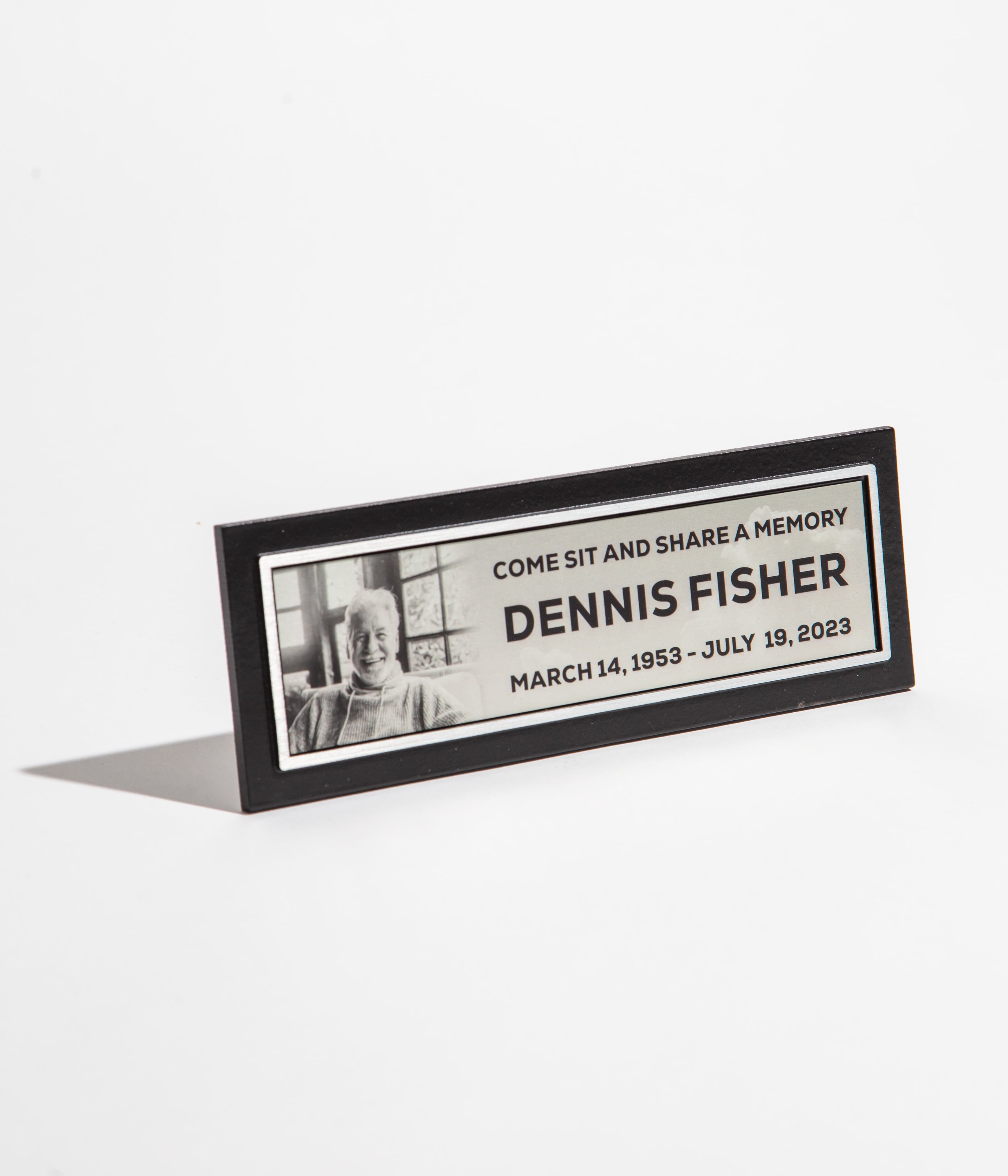 Memorial plaque with a photo and text commemorating Dennis Fisher, dated March 14, 1953 - July 19, 2023.