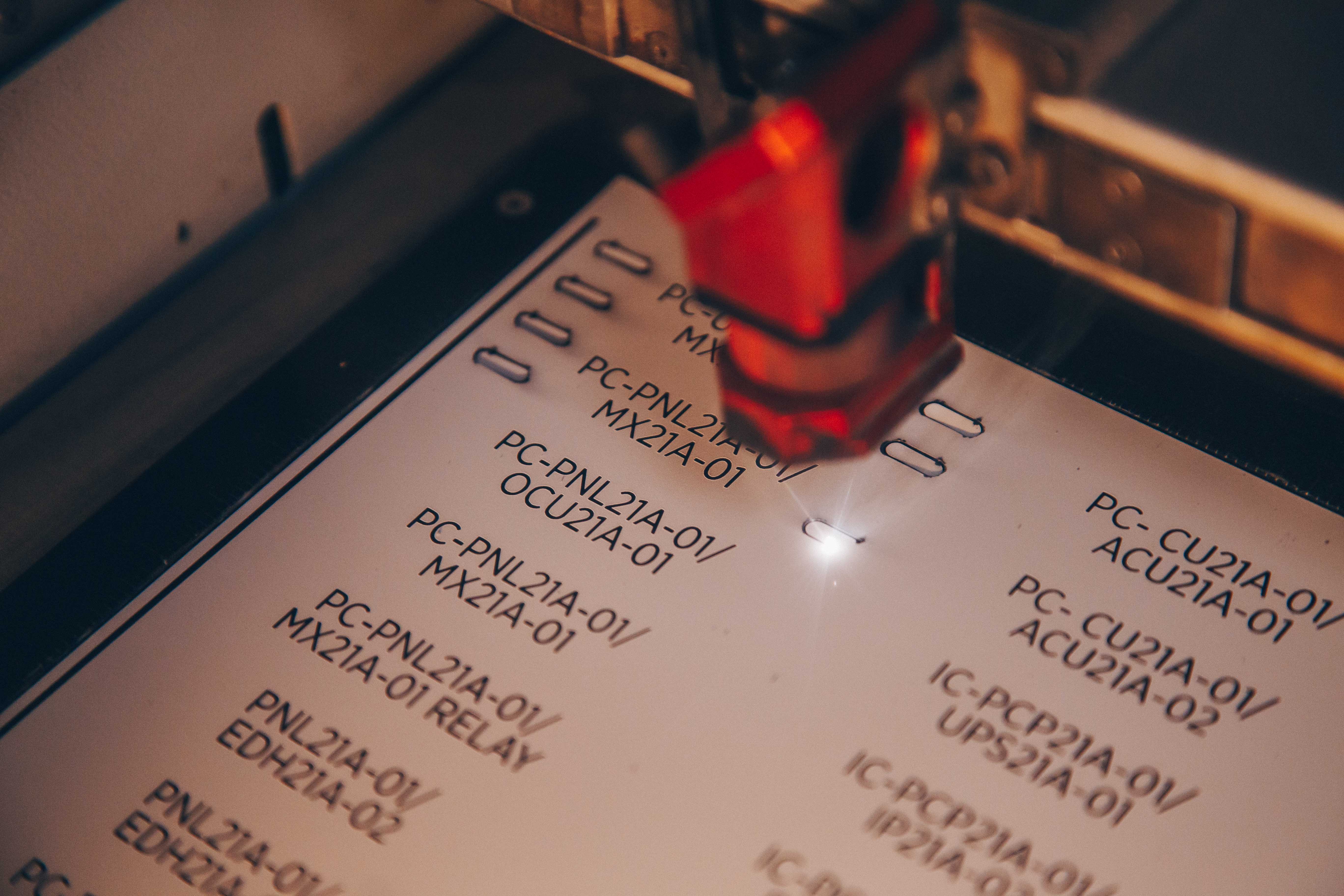Close-up photo of an engraved label . A red light or laser pointer is visible in the frame, highlighting one of the codes. The image has a shallow depth of field with some text appearing slightly blurred.
