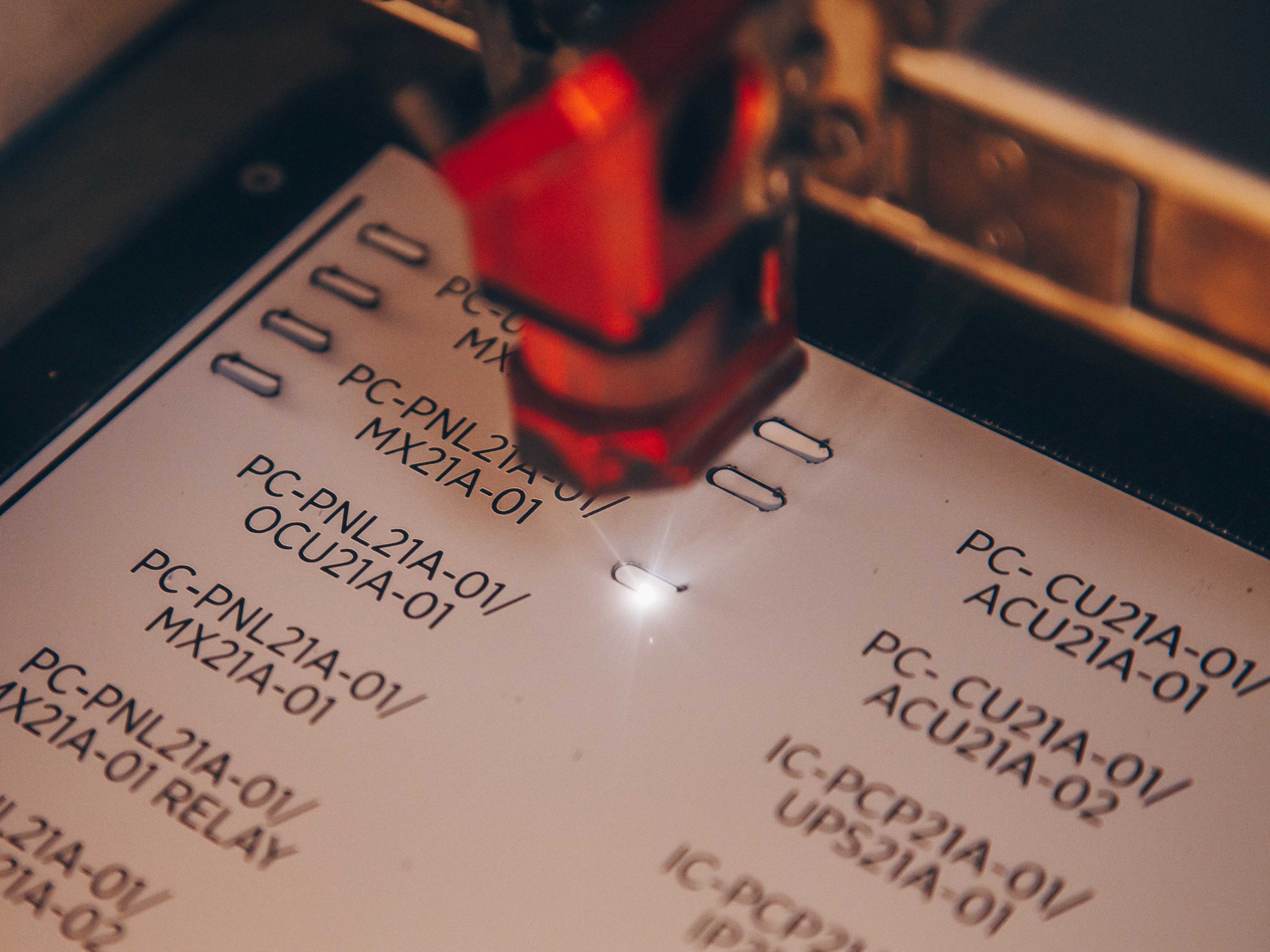 Close-up photo of an engraved label . A red light or laser pointer is visible in the frame, highlighting one of the codes. The image has a shallow depth of field with some text appearing slightly blurred.