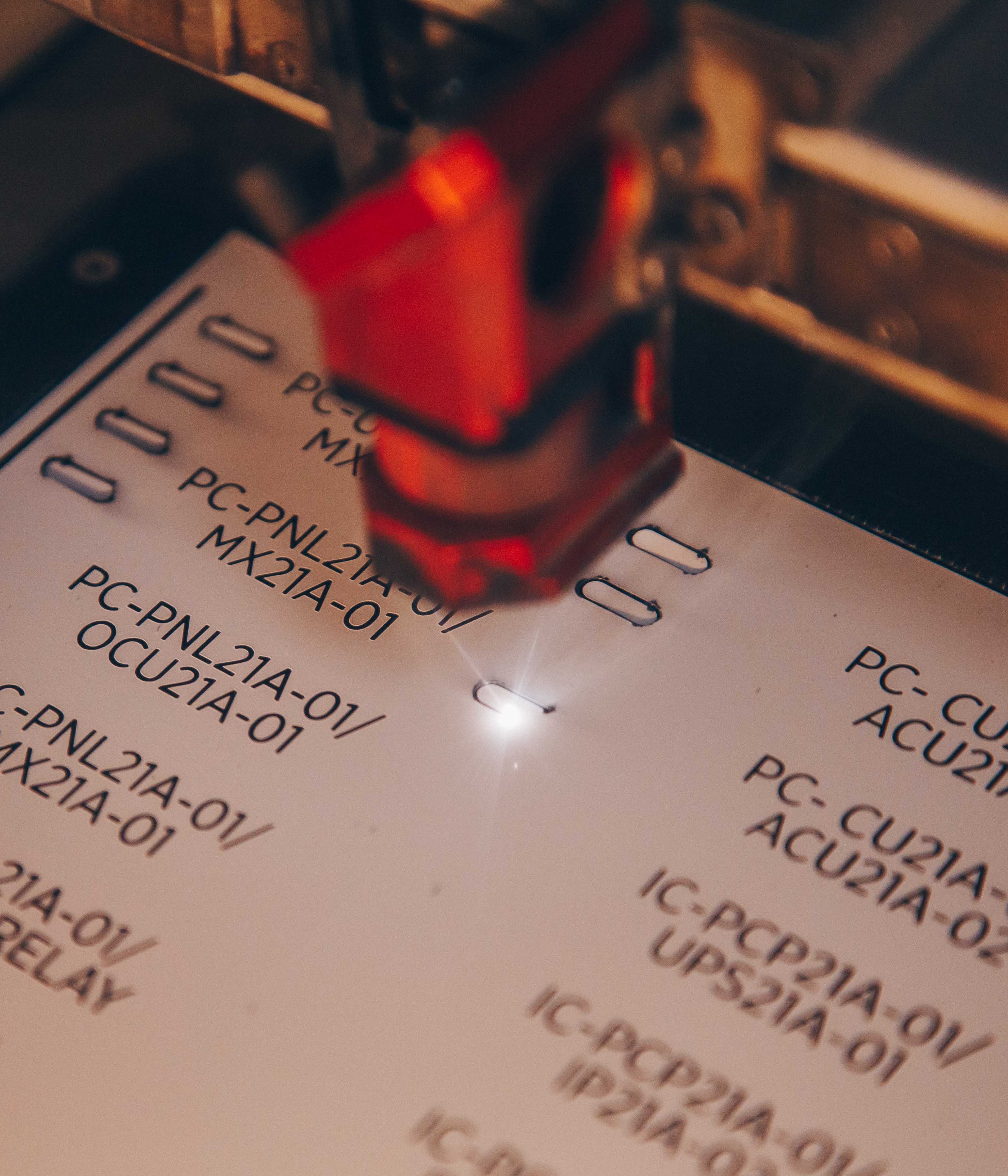 Close-up photo of an engraved label . A red light or laser pointer is visible in the frame, highlighting one of the codes. The image has a shallow depth of field with some text appearing slightly blurred.