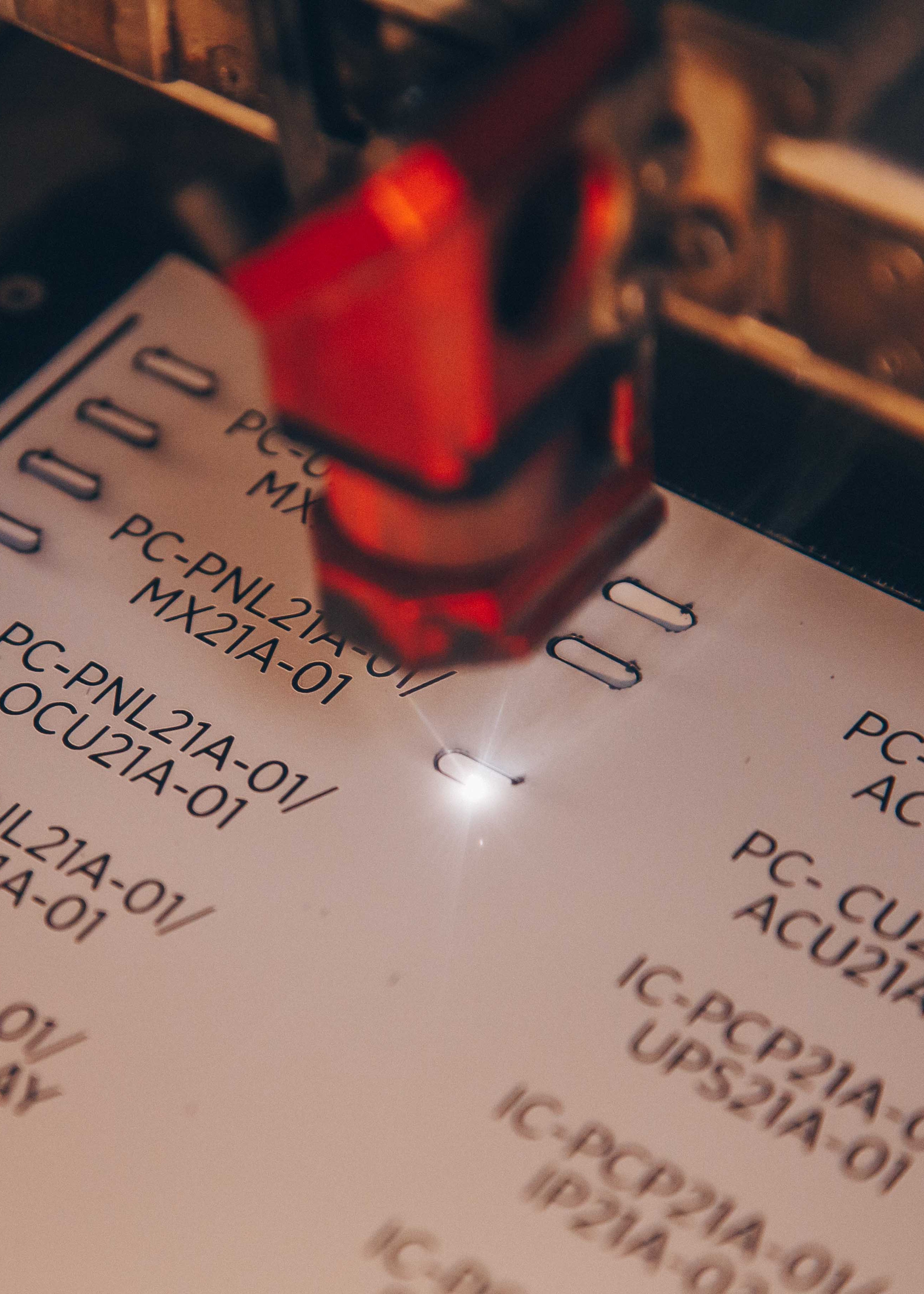 Close-up photo of an engraved label . A red light or laser pointer is visible in the frame, highlighting one of the codes. The image has a shallow depth of field with some text appearing slightly blurred.