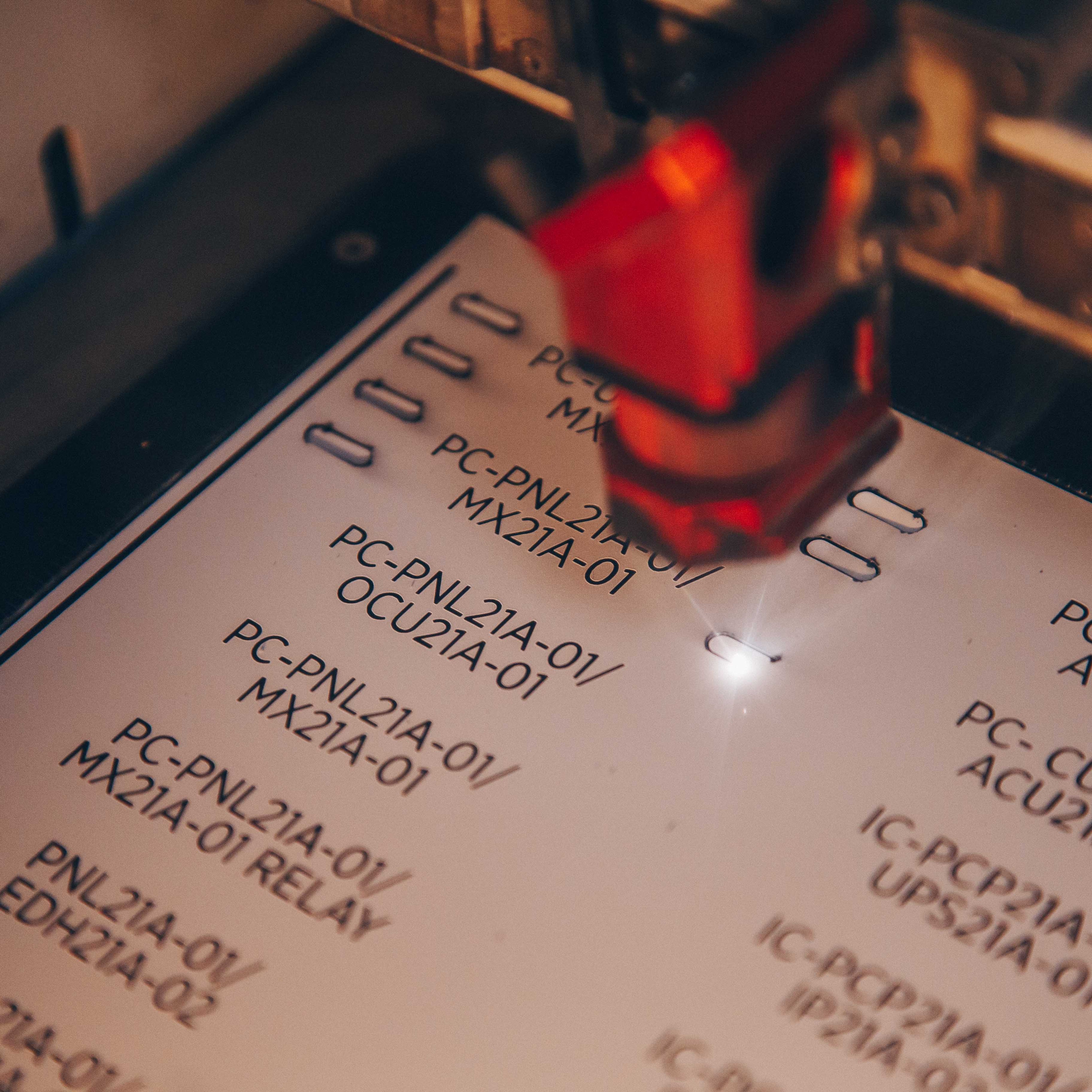 Close-up photo of an engraved label . A red light or laser pointer is visible in the frame, highlighting one of the codes. The image has a shallow depth of field with some text appearing slightly blurred.
