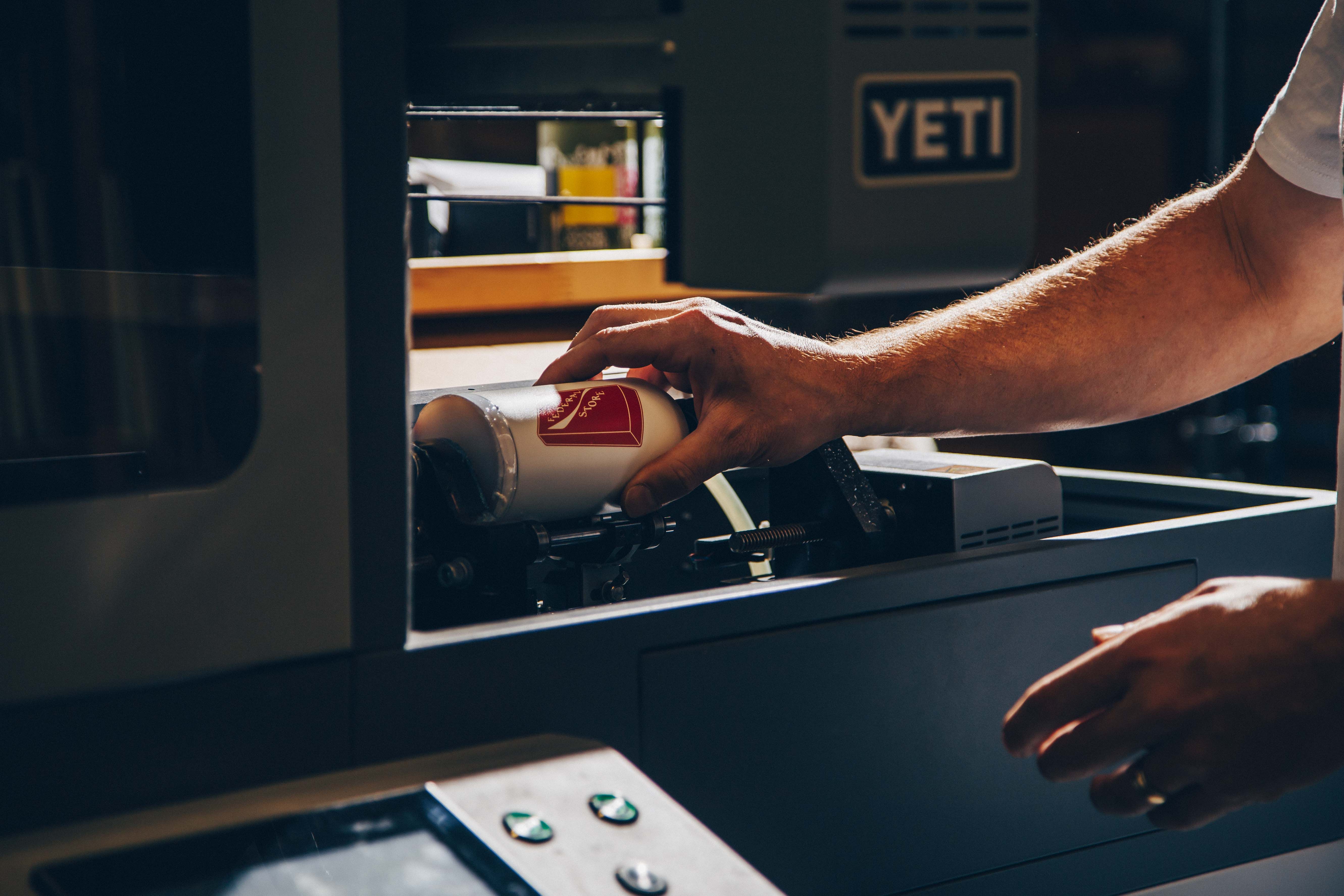 A hand positions a white cylindrical drinkware item inside a Yeti rotary printer, showing the precise placement for custom printing.