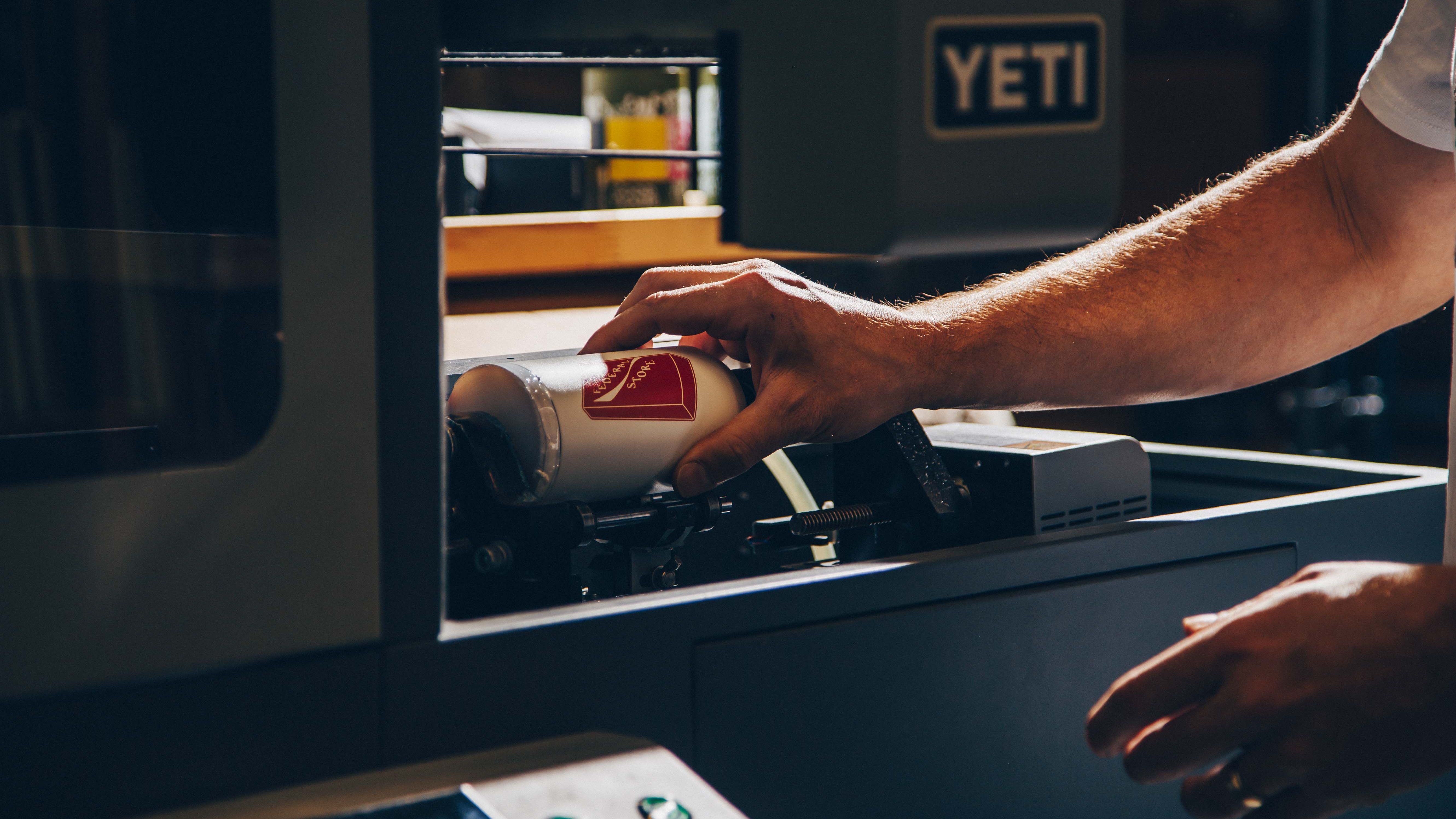 A hand positions a white cylindrical drinkware item inside a Yeti rotary printer, showing the precise placement for custom printing.