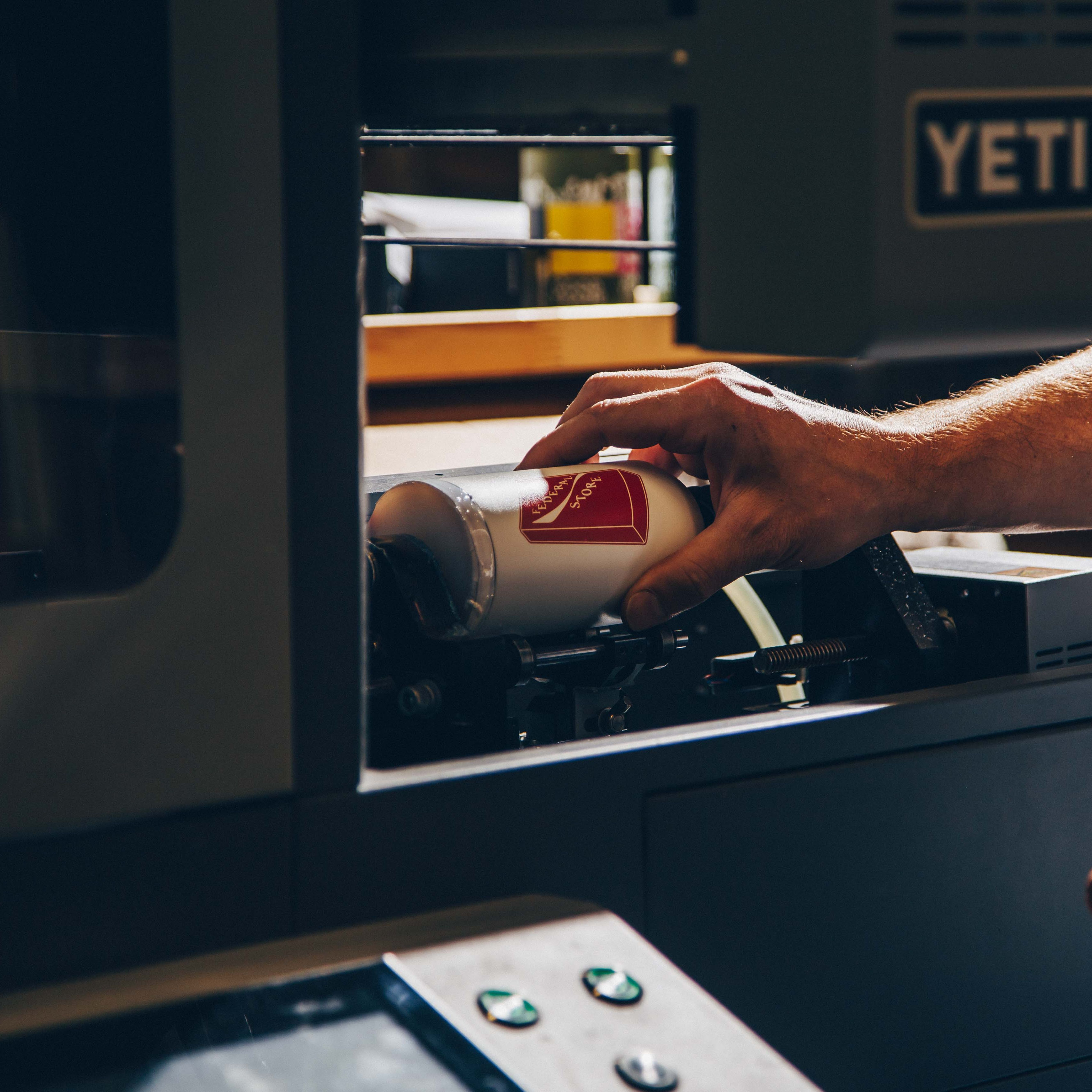 A hand positions a white cylindrical drinkware item inside a Yeti rotary printer, showing the precise placement for custom printing.