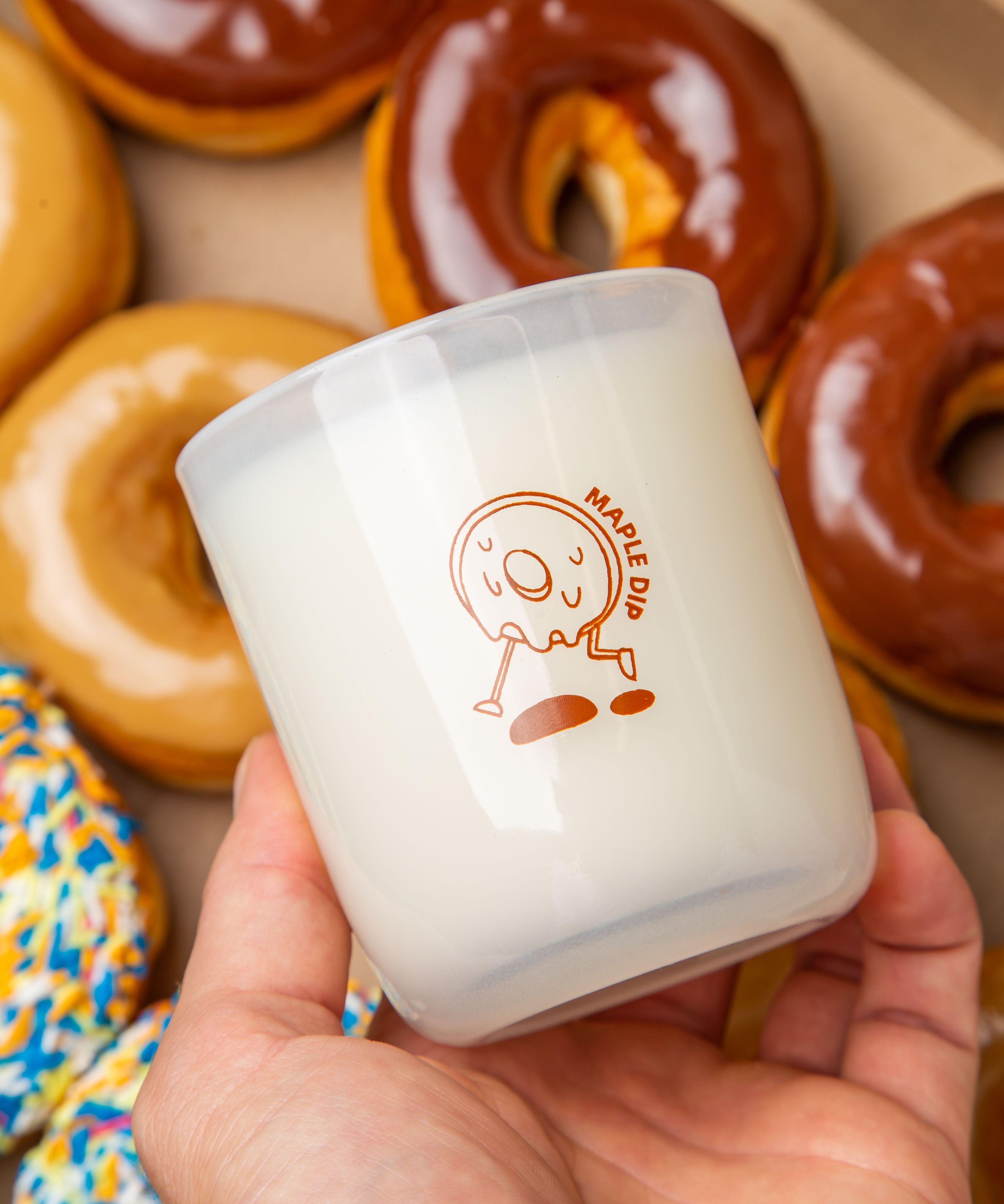 Hand holding a Milk Jar Candle Co. branded candle with a maple dip donut design, surrounded by glazed donuts.