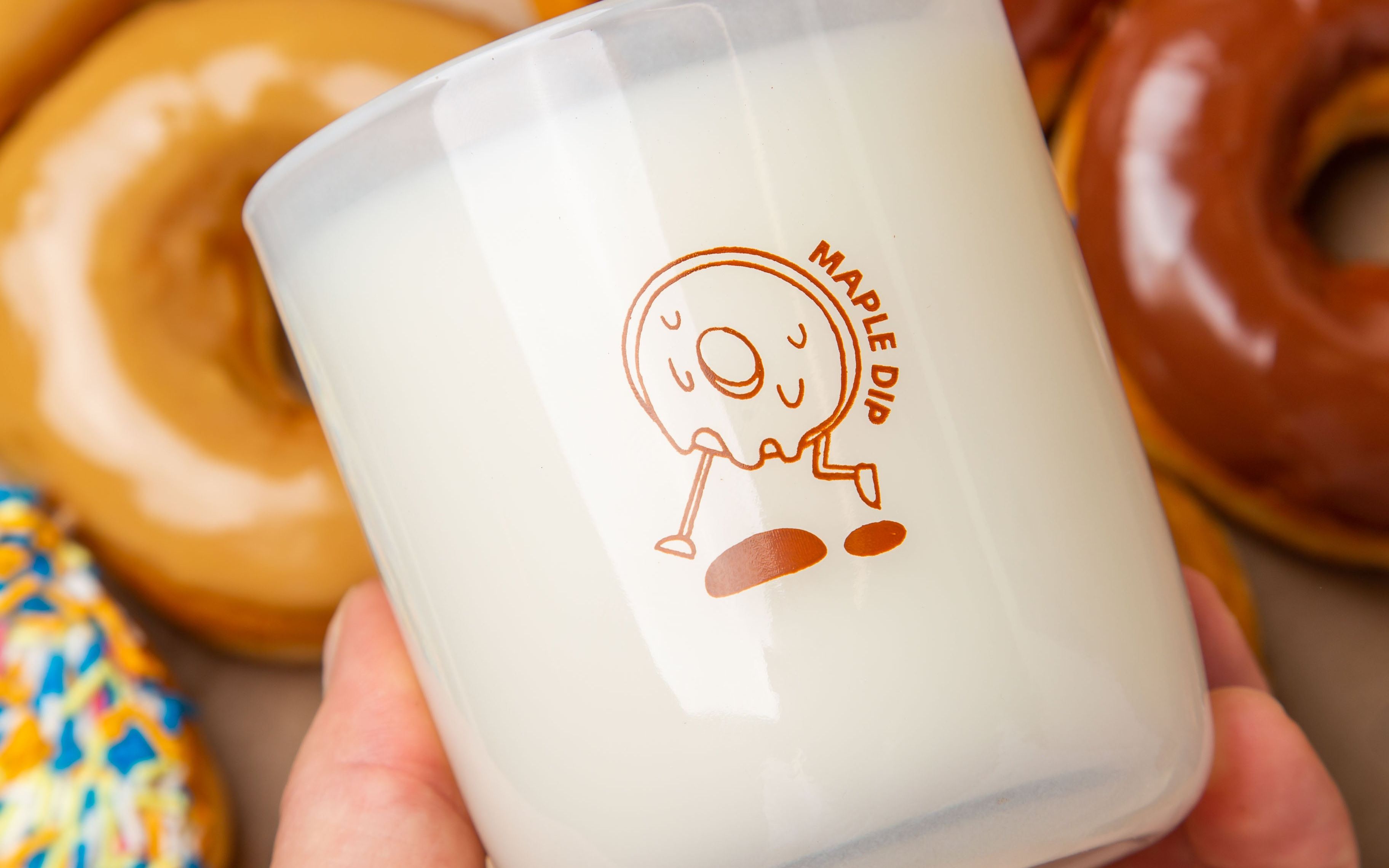 Hand holding a Milk Jar Candle Co. branded candle with a maple dip donut design, surrounded by glazed donuts.