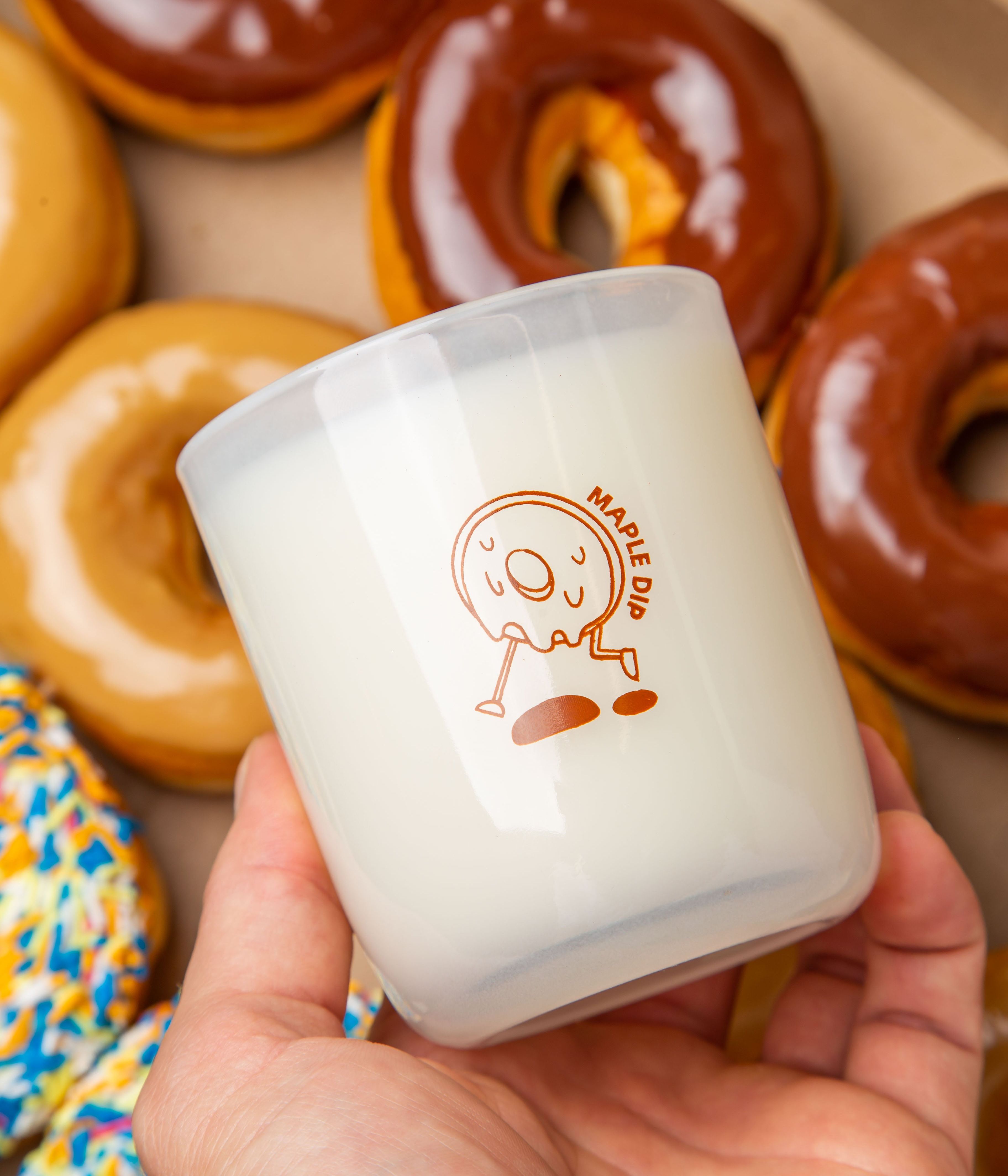 Hand holding a Milk Jar Candle Co. branded candle with a maple dip donut design, surrounded by glazed donuts.