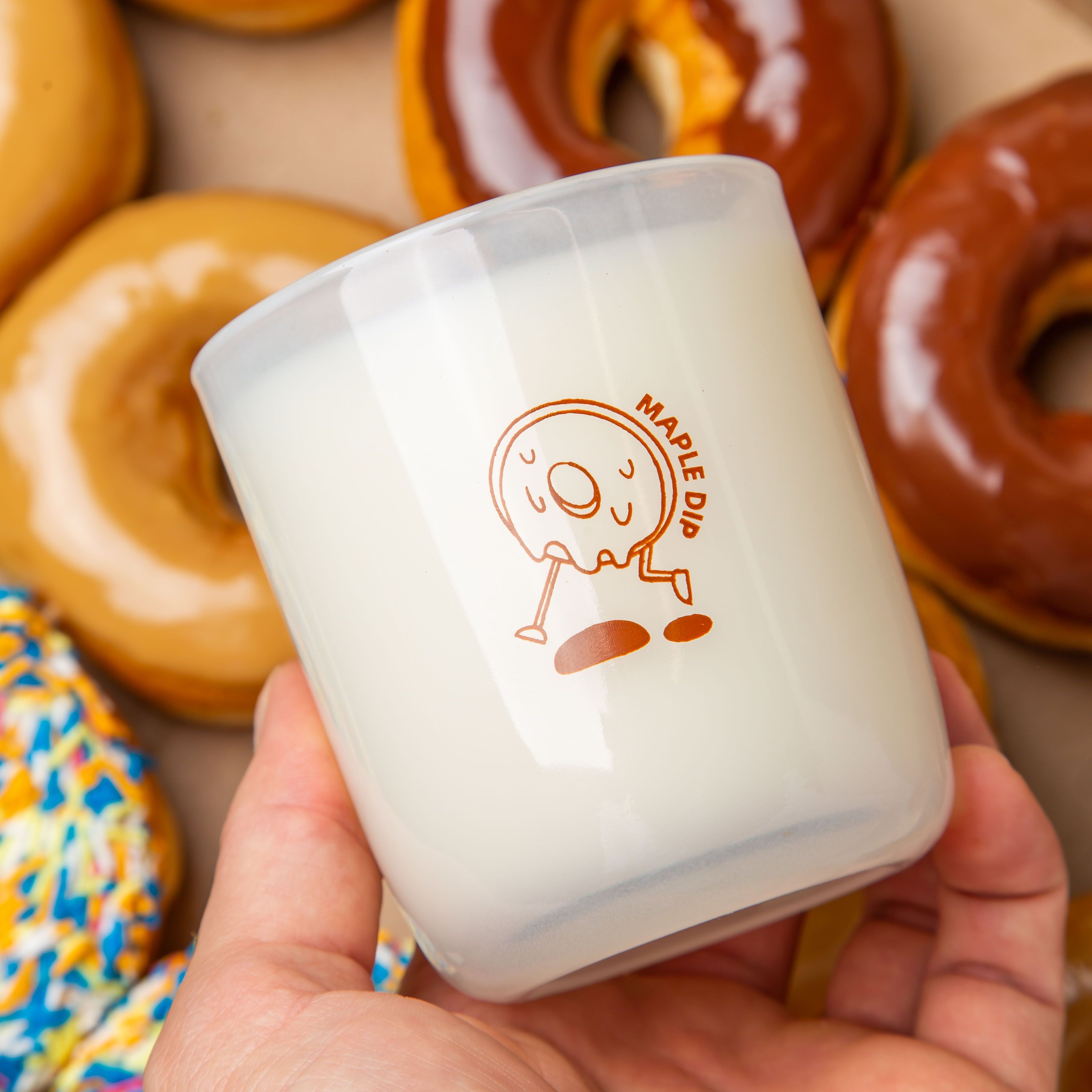 Hand holding a Milk Jar Candle Co. branded candle with a maple dip donut design, surrounded by glazed donuts.