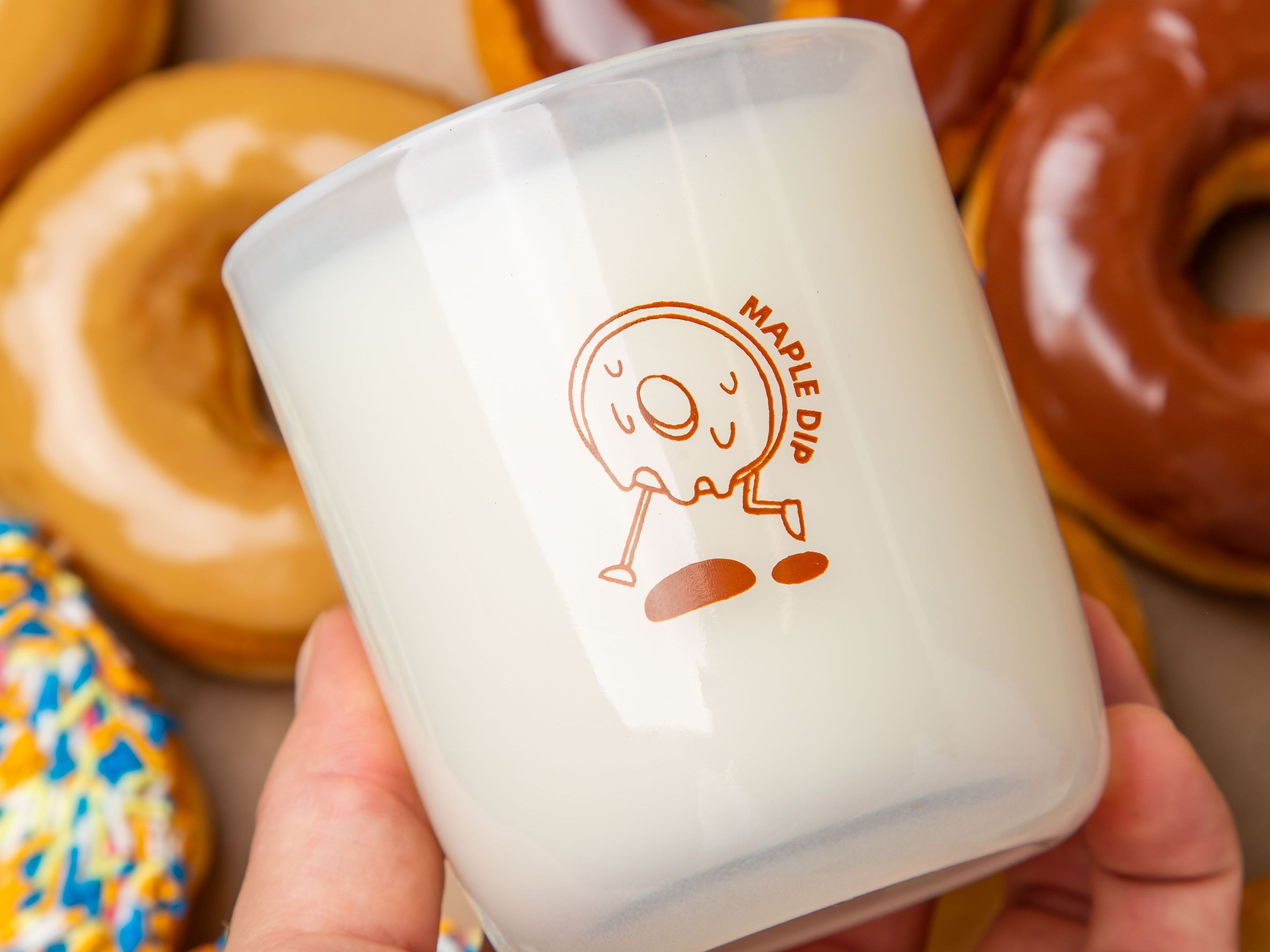 Hand holding a Milk Jar Candle Co. branded candle with a maple dip donut design, surrounded by glazed donuts.