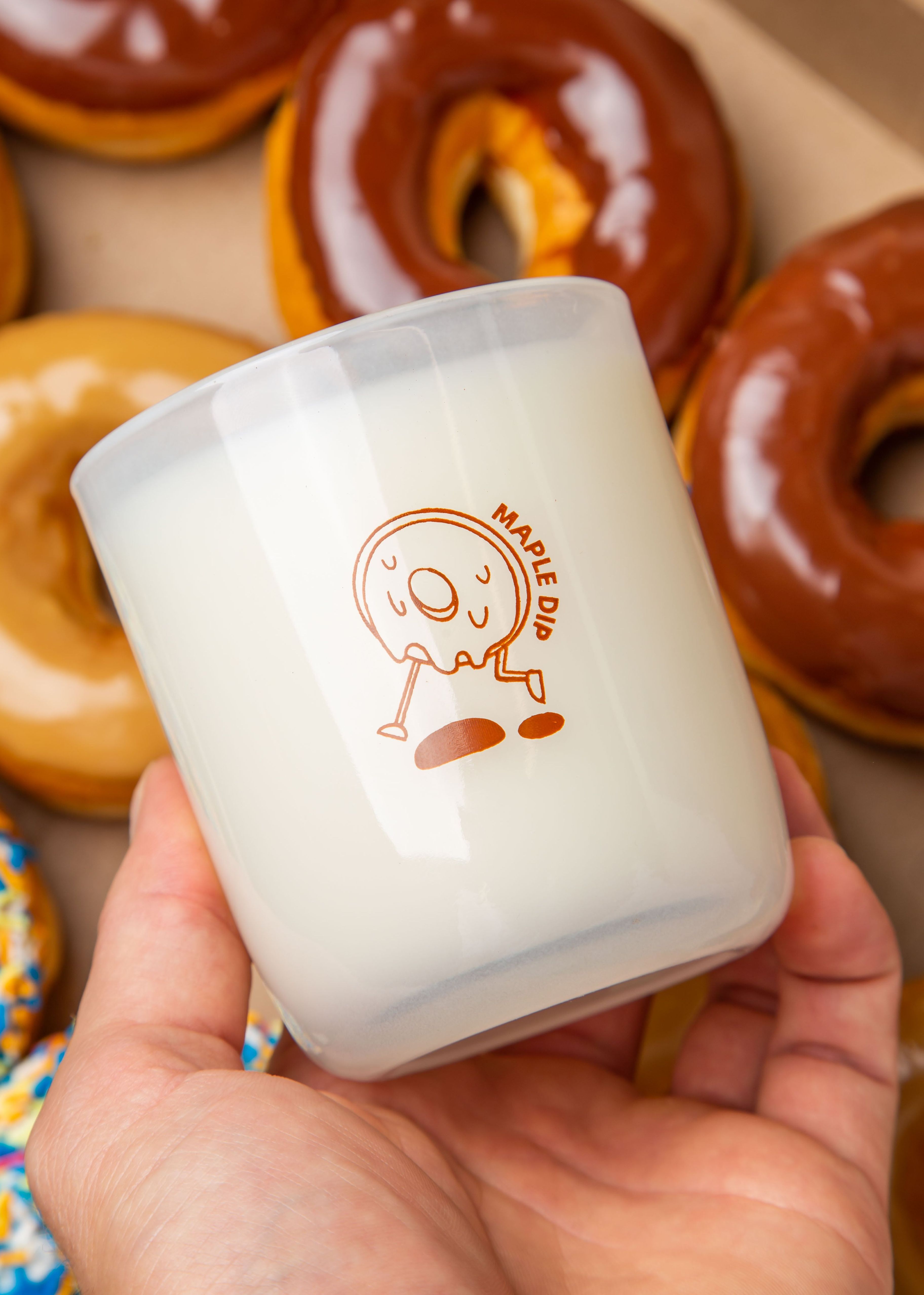 Hand holding a Milk Jar Candle Co. branded candle with a maple dip donut design, surrounded by glazed donuts.