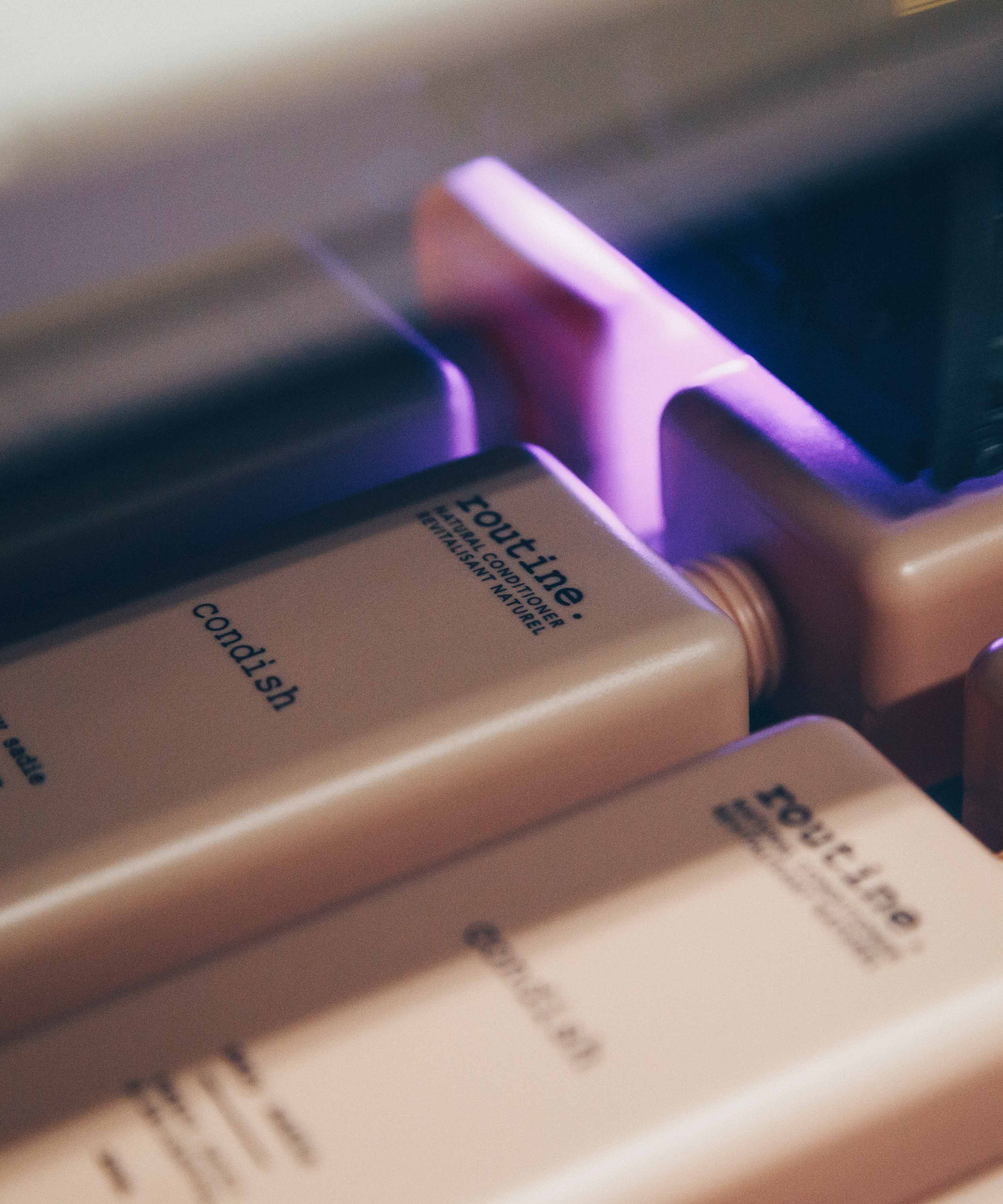 A close-up view of several rectangular bottles, likely containing hair conditioner, arranged in a row. The bottles are a soft beige color with black text, featuring the brand name 'routine' and the product type 'condish'. The image captures a moment in a manufacturing or packaging process, as indicated by the presence of machinery above the bottles, emitting a soft purple light.