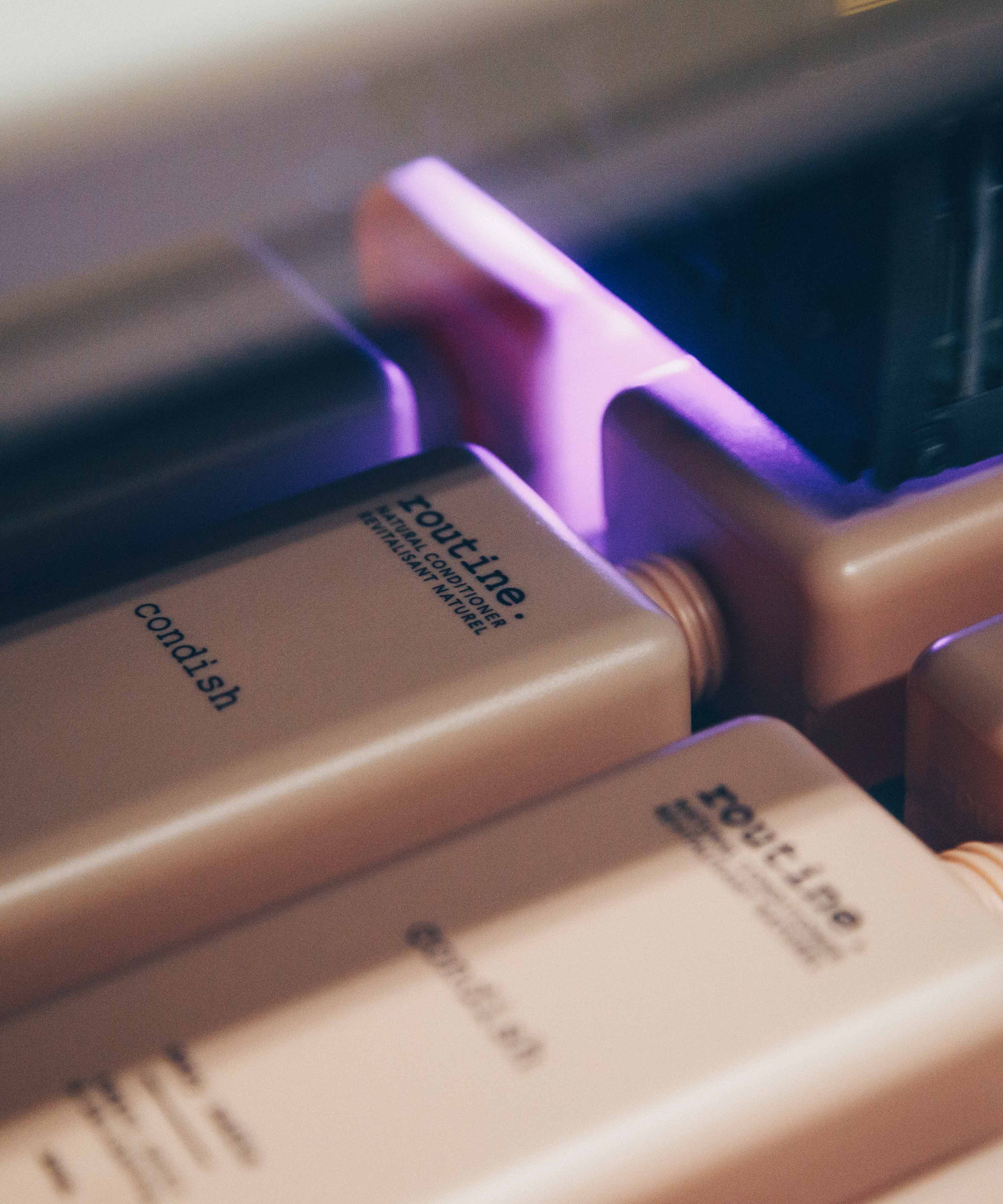 A close-up view of several rectangular bottles, likely containing hair conditioner, arranged in a row. The bottles are a soft beige color with black text, featuring the brand name 'routine' and the product type 'condish'. The image captures a moment in a manufacturing or packaging process, as indicated by the presence of machinery above the bottles, emitting a soft purple light.