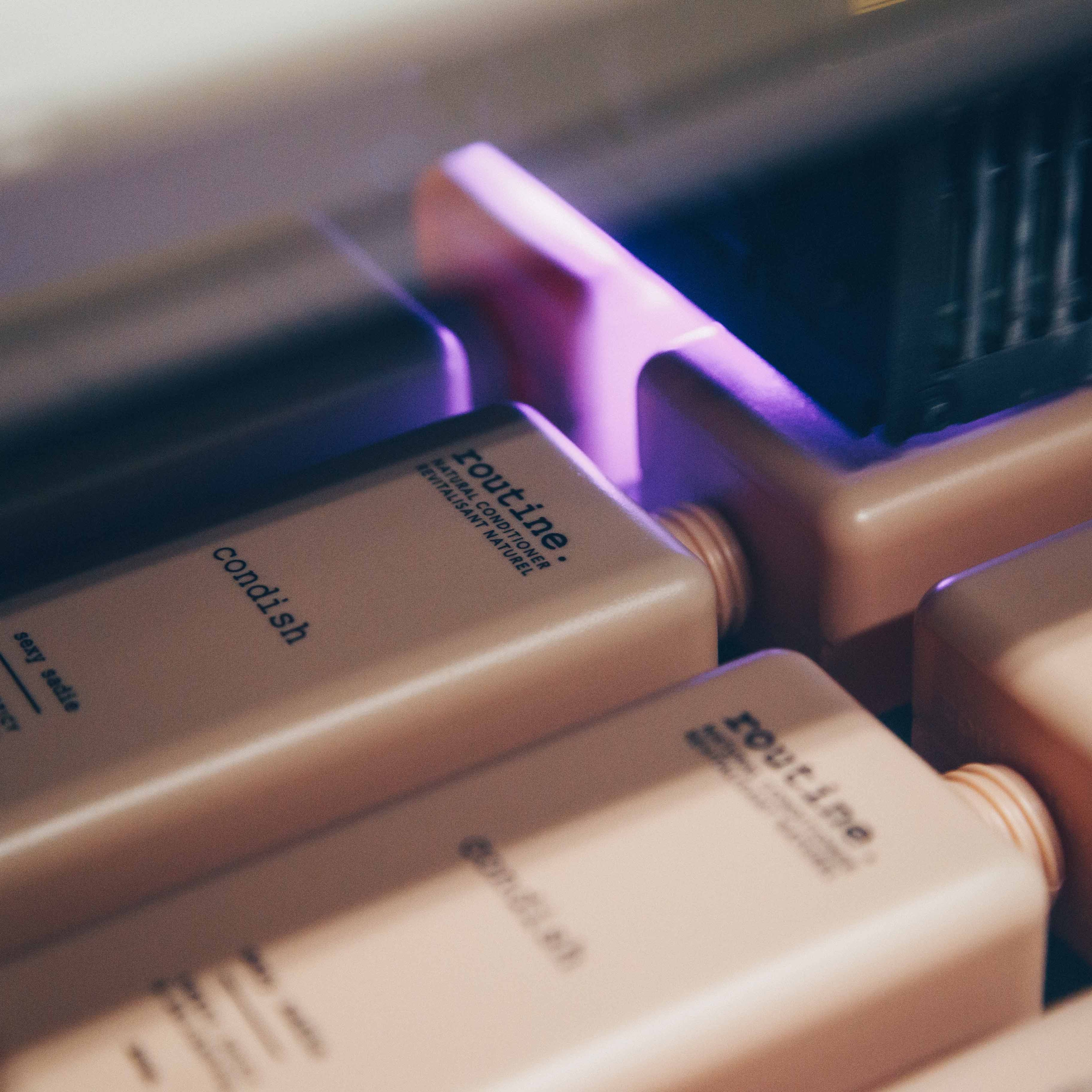 A close-up view of several rectangular bottles, likely containing hair conditioner, arranged in a row. The bottles are a soft beige color with black text, featuring the brand name 'routine' and the product type 'condish'. The image captures a moment in a manufacturing or packaging process, as indicated by the presence of machinery above the bottles, emitting a soft purple light.