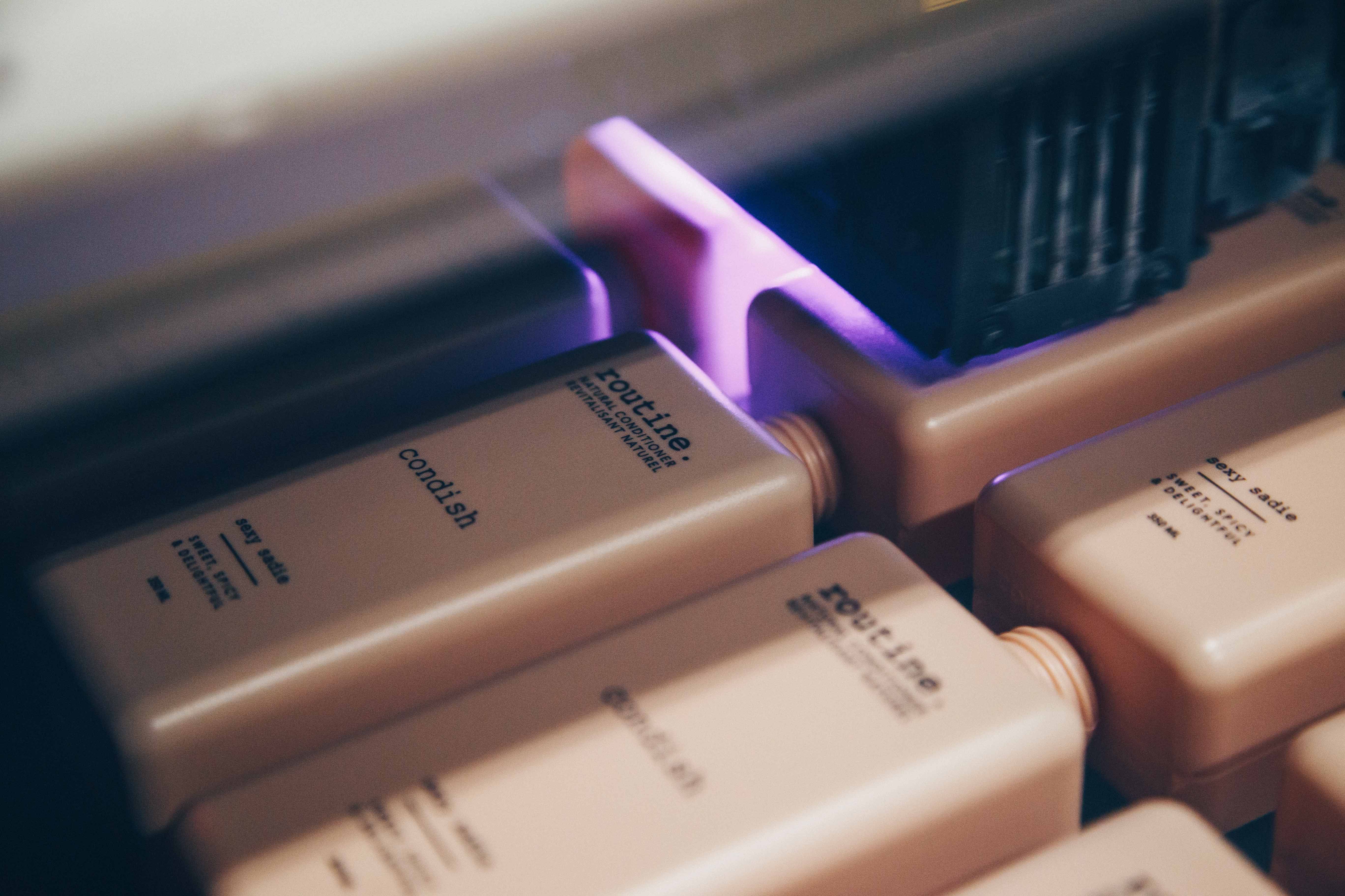 A close-up view of several rectangular bottles, likely containing hair conditioner, arranged in a row. The bottles are a soft beige color with black text, featuring the brand name 'routine' and the product type 'condish'. The image captures a moment in a manufacturing or packaging process, as indicated by the presence of machinery above the bottles, emitting a soft purple light.