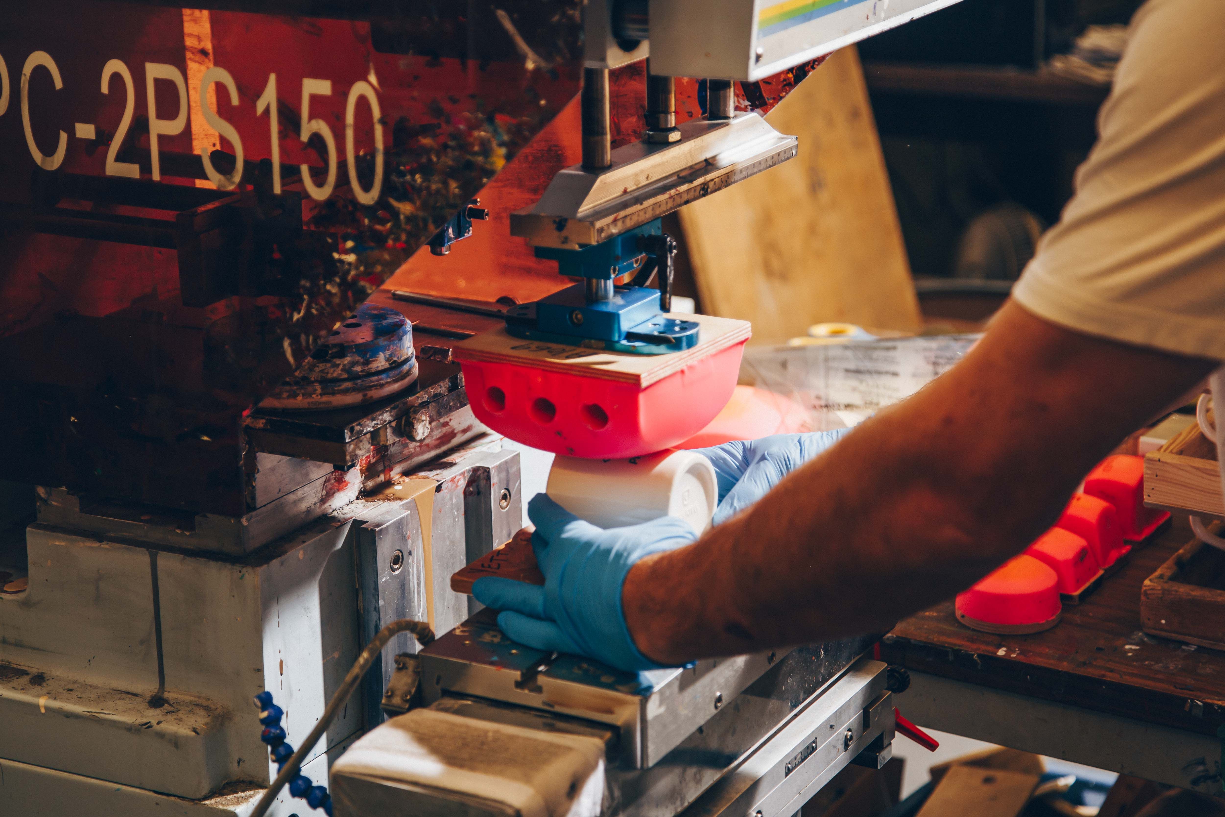 A pad printing machine in operation, labeled "MPC-2PS150," with a worker positioning a ceramic mug under a red silicone pad for branding. The industrial setup highlights precision printing for promotional products.