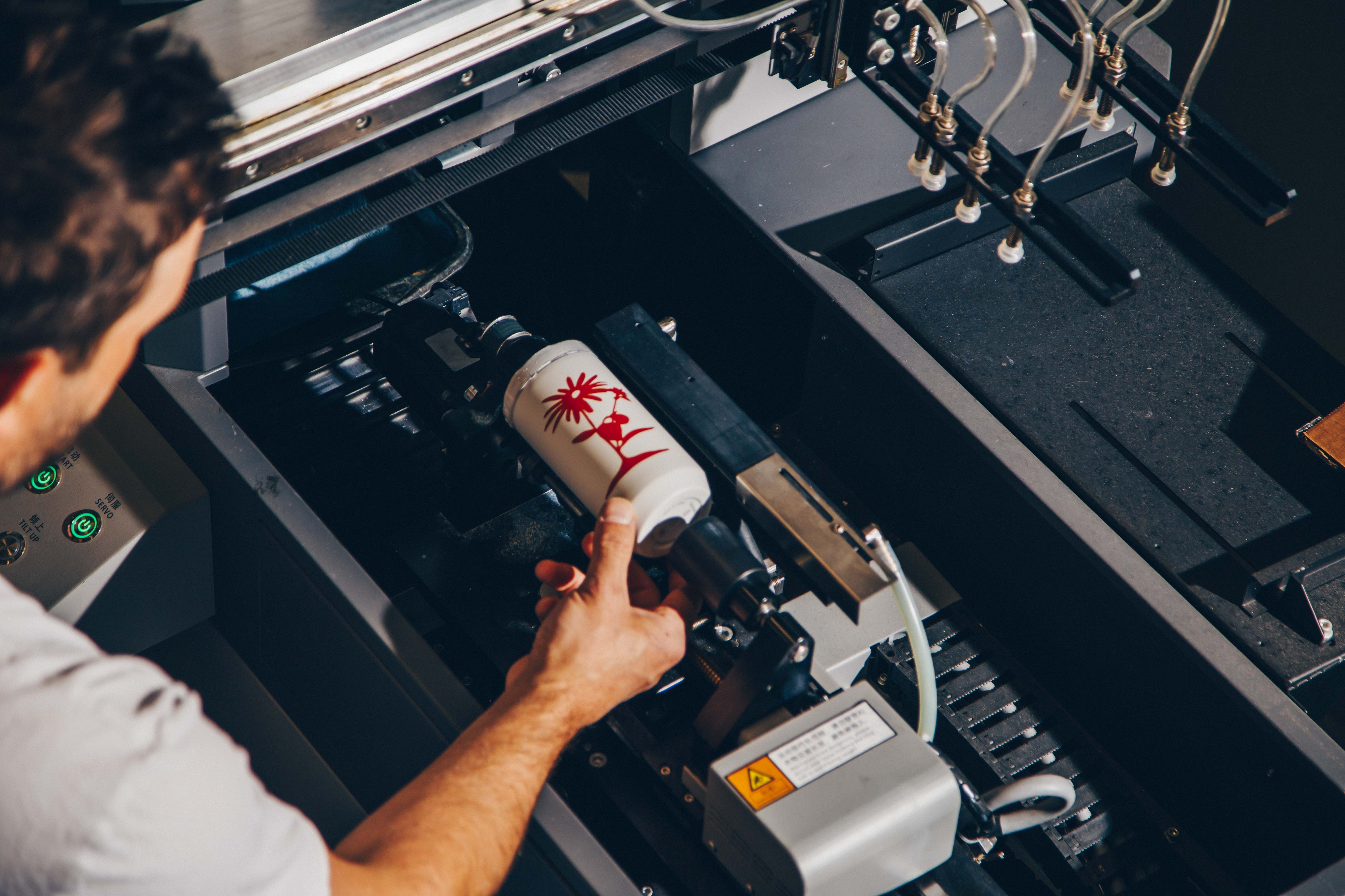 A person adjusts a white cylindrical bottle within a digital rotary printer, showcasing a custom red design being applied to the surface.