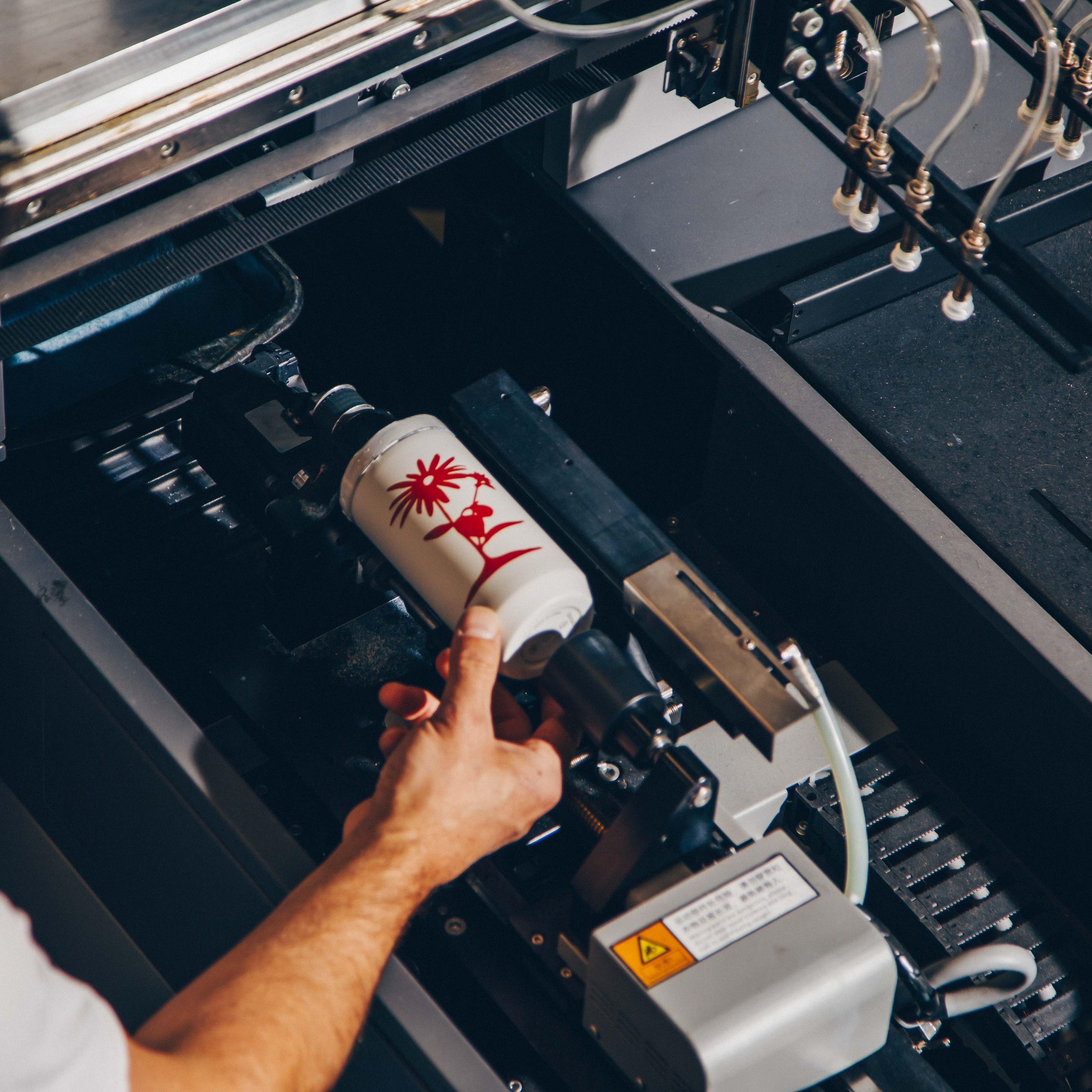 A person adjusts a white cylindrical bottle within a digital rotary printer, showcasing a custom red design being applied to the surface.