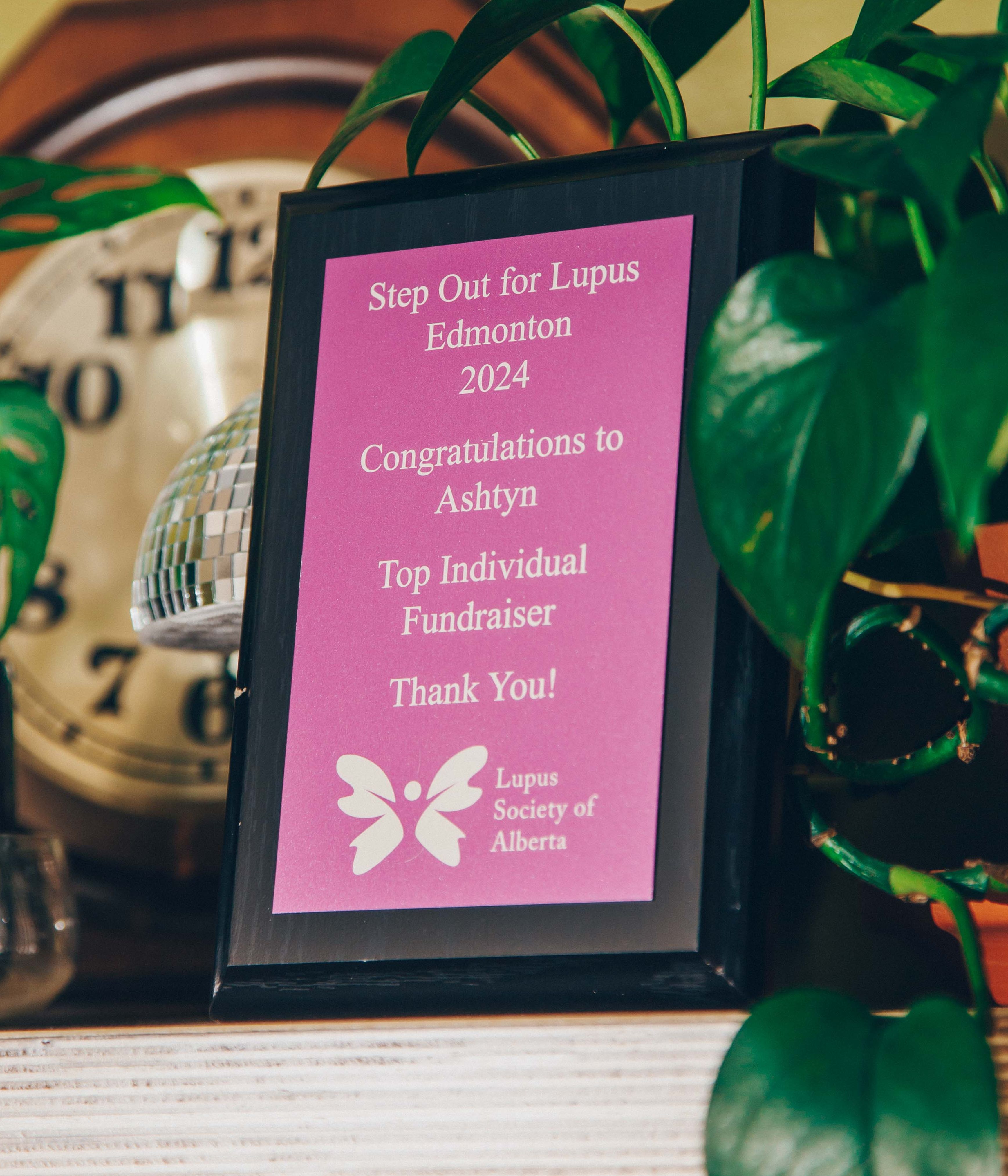 Award plaque for Step Out for Lupus Edmonton 2024, congratulating Ashtyn as top individual fundraiser, surrounded by plants and a clock.