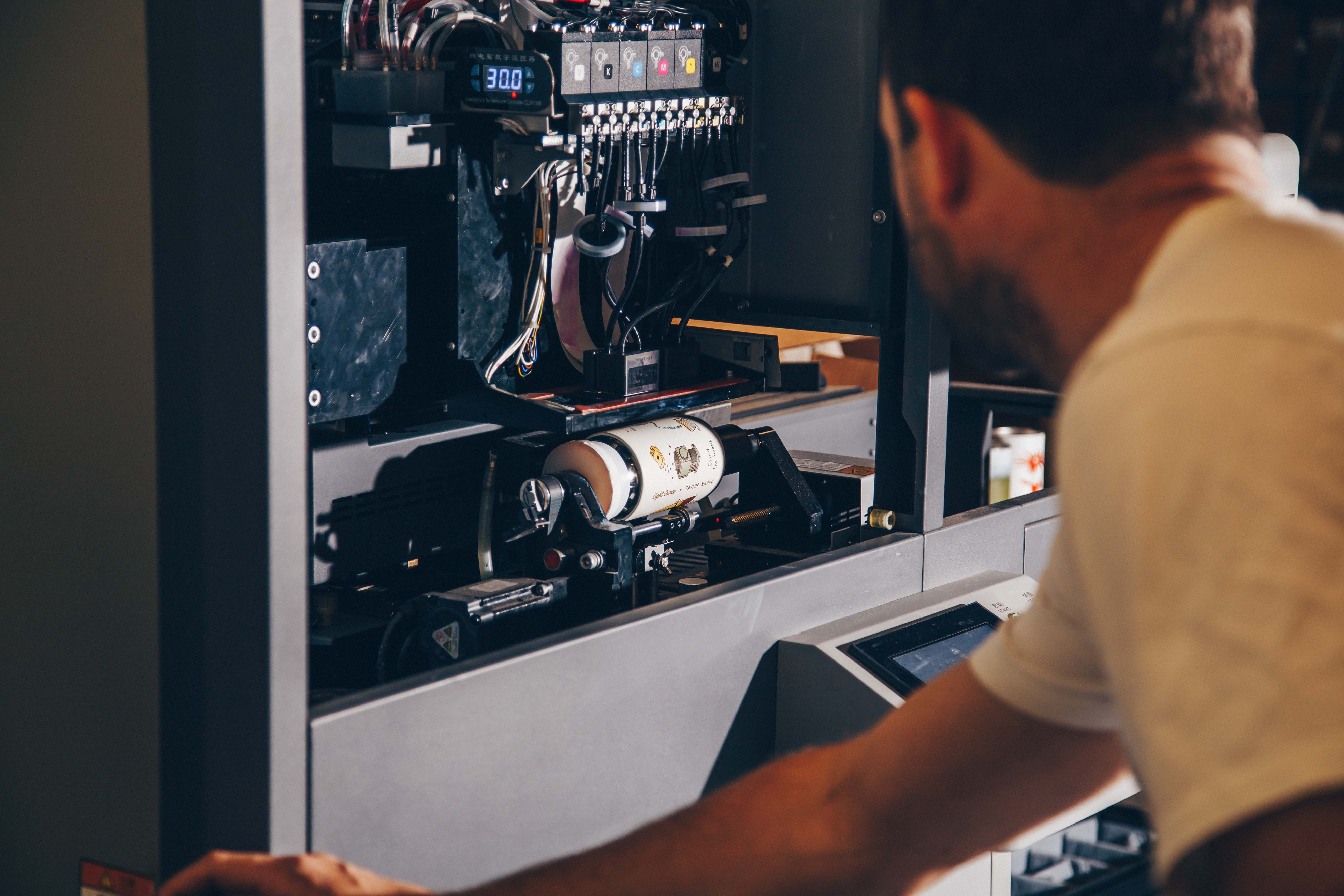 A person supervises the operation of a rotary printer, observing the design application on a bottle in real time.